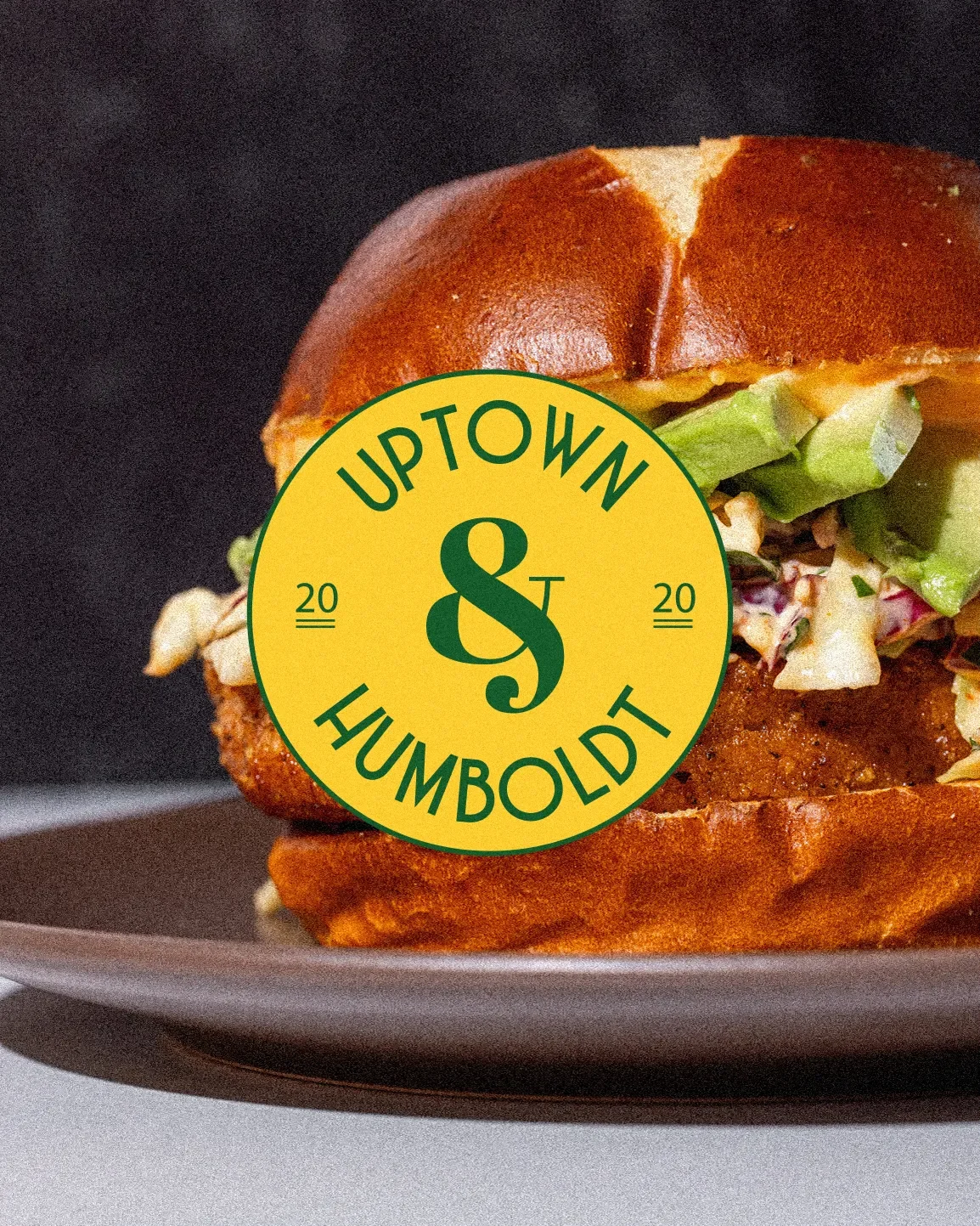 Close-up of a sandwich with lettuce, coleslaw, and fried chicken on a bun, placed on a black plate, with a green and yellow Uptown & Humboldt logo overlay in the center.