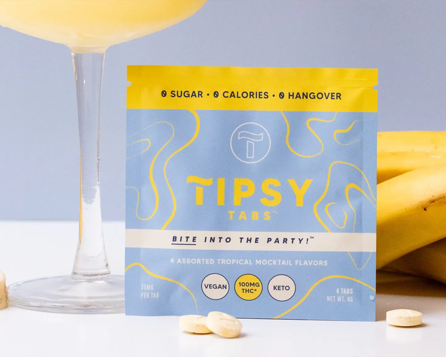 A box of Tipsy Tabs tropical mocktail flavor, labeled as vegan, keto, containing 100mg THC, with four tablets visible around the box, partially obscured bananas in the background, and a glass with a drink to the left.