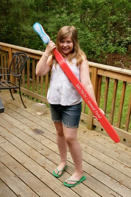 Photo of a girl holding a giant red toothbrush with the words “Brown Annual Fund” on the handle to show two important habits, brushing your teeth and giving through the annual fund.
