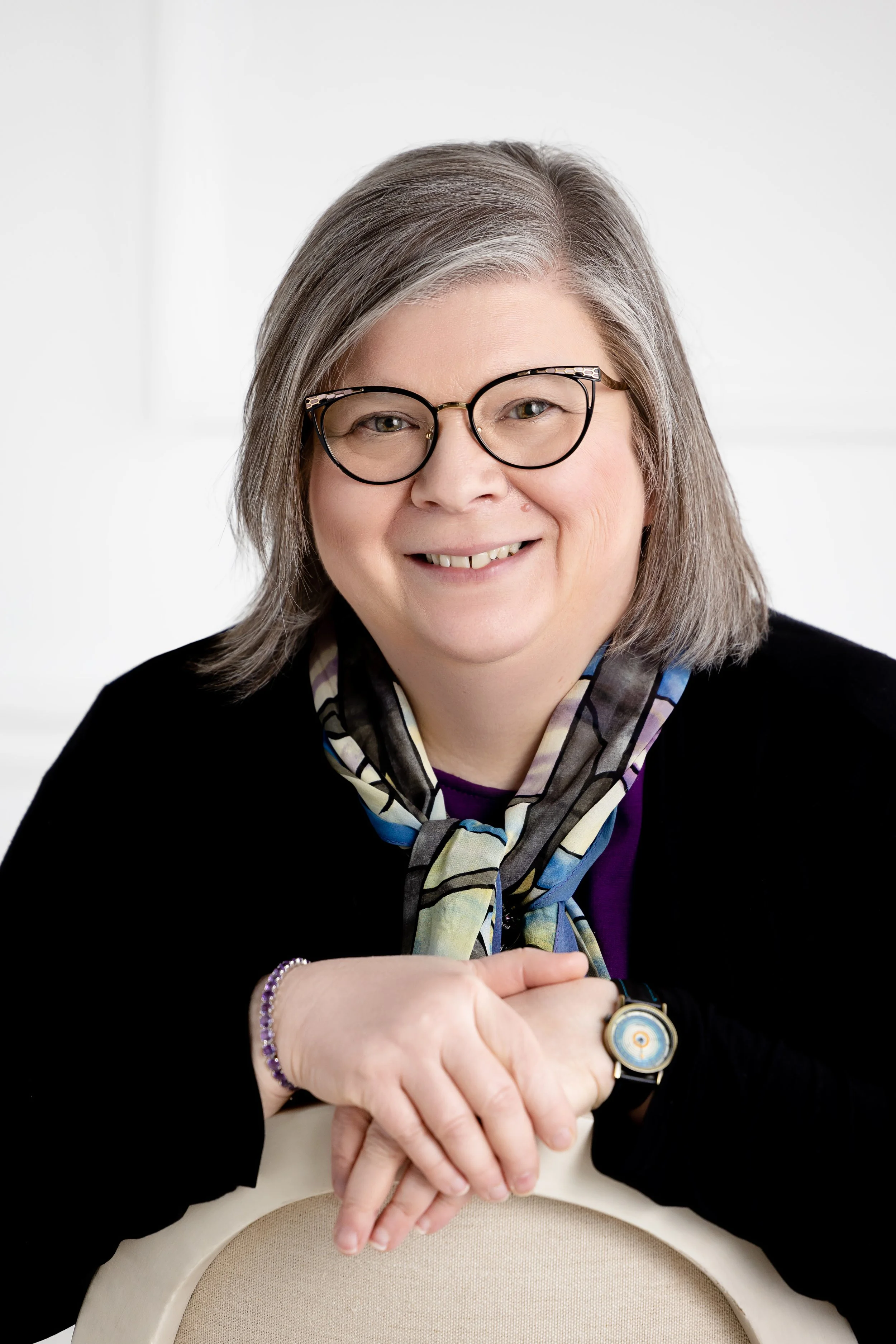 Photo of Tammie Ruda, founder. She is smiling, wearing glasses, a black sweater and a colorful scarf, and posing with her hands crossed.