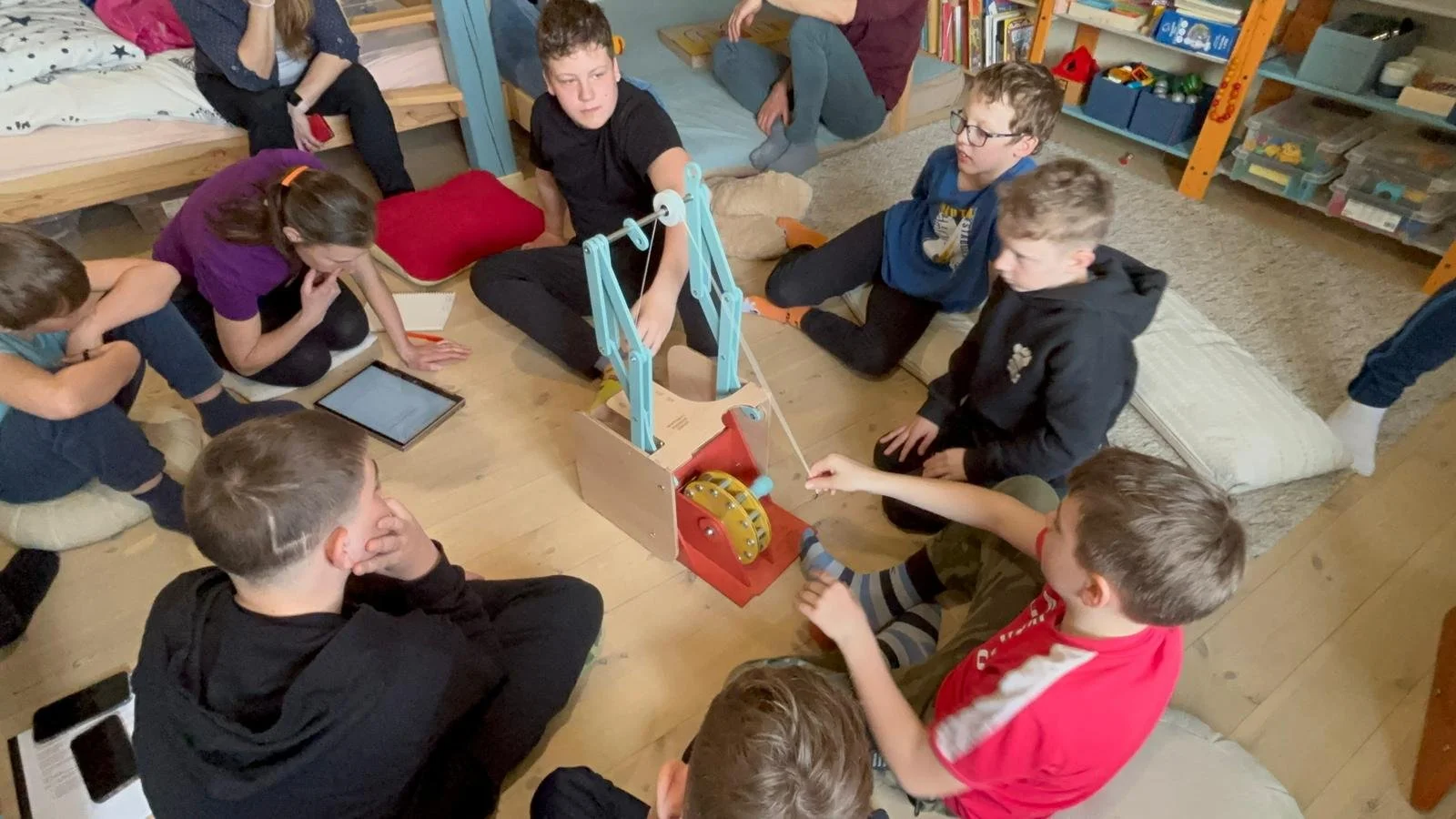 Group of children gathered around a wooden mechanical device on the floor, engaging in discussion or activity in a room with shelves and books.
