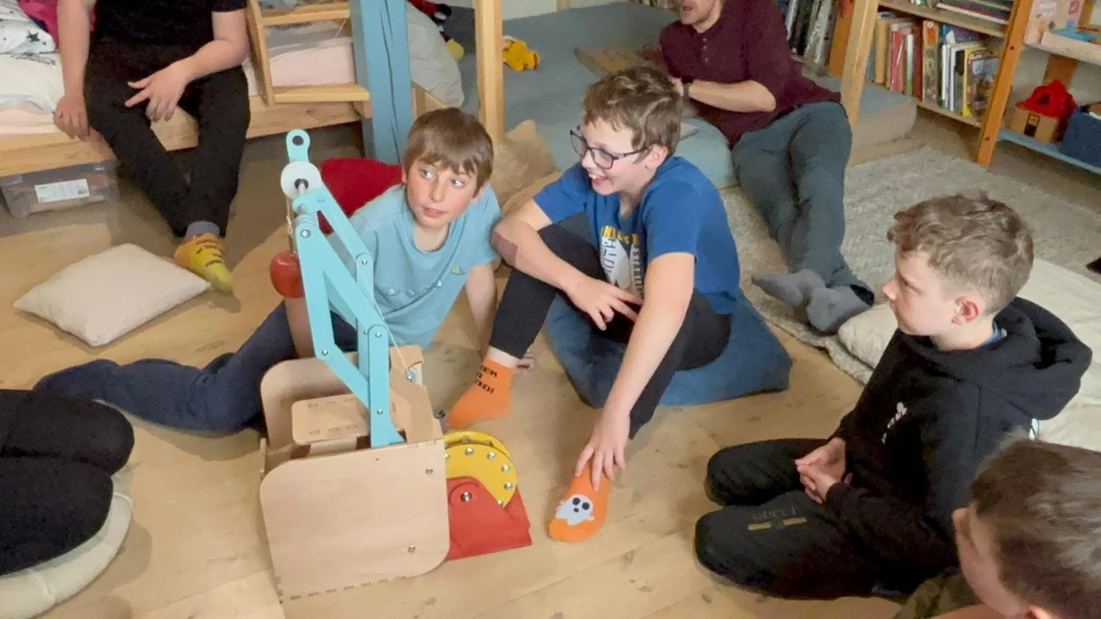 Children sitting on the floor engaged with a wooden mechanical contraption in a room with shelves and cushions.