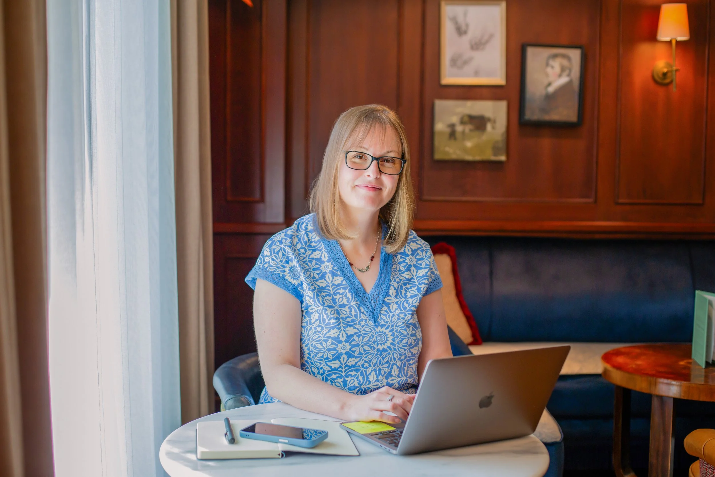 A woman with glasses and shoulder-length blonde hair, wearing a blue patterned blouse, sitting at a round table with a laptop, phone, pen, and notebook, in a cozy room with wooden walls and framed artwork.