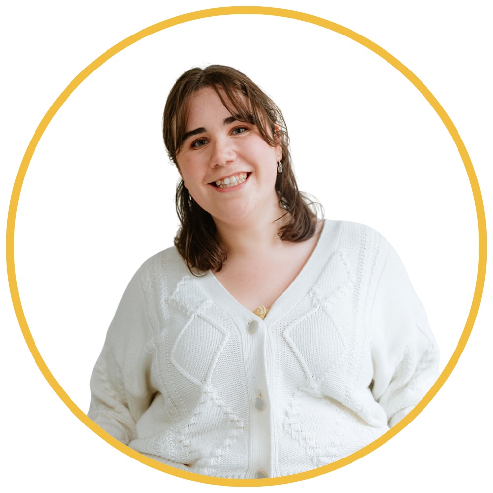 A young woman with brown hair and fair skin smiling at the camera, wearing a white knitted sweater, against a white background with a yellow circular frame.