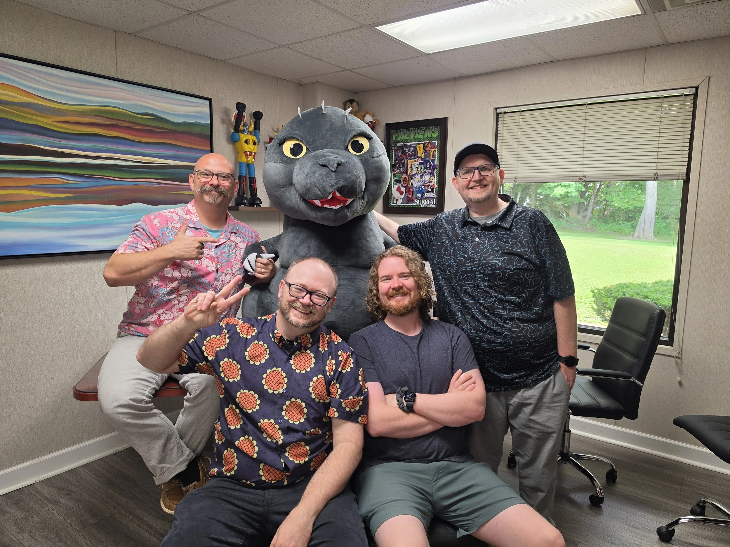 Five people posing with a person in a large gray cat costume inside a room with artwork and a window showing green trees outside.