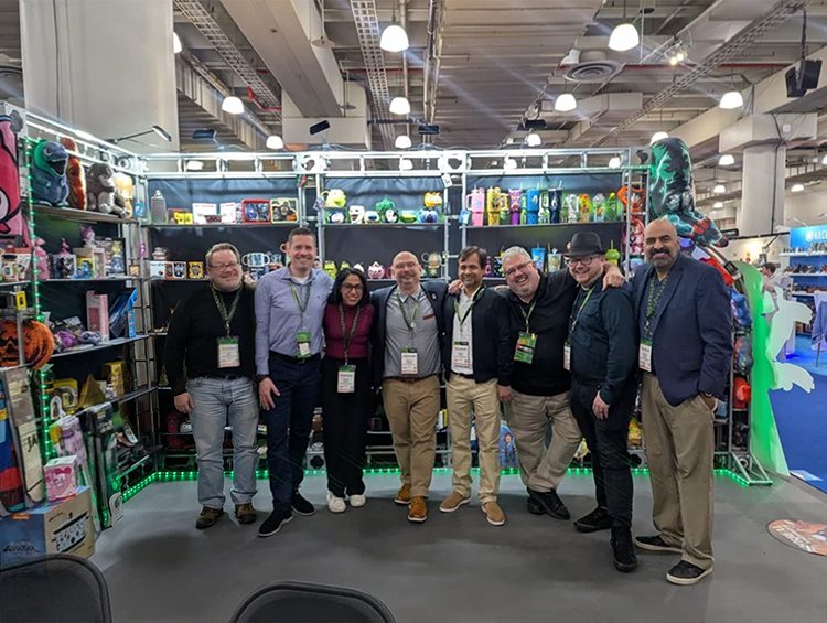 Group of eight people standing in front of a toy booth at a trade show, smiling, with shelves of toys and plush characters behind them.