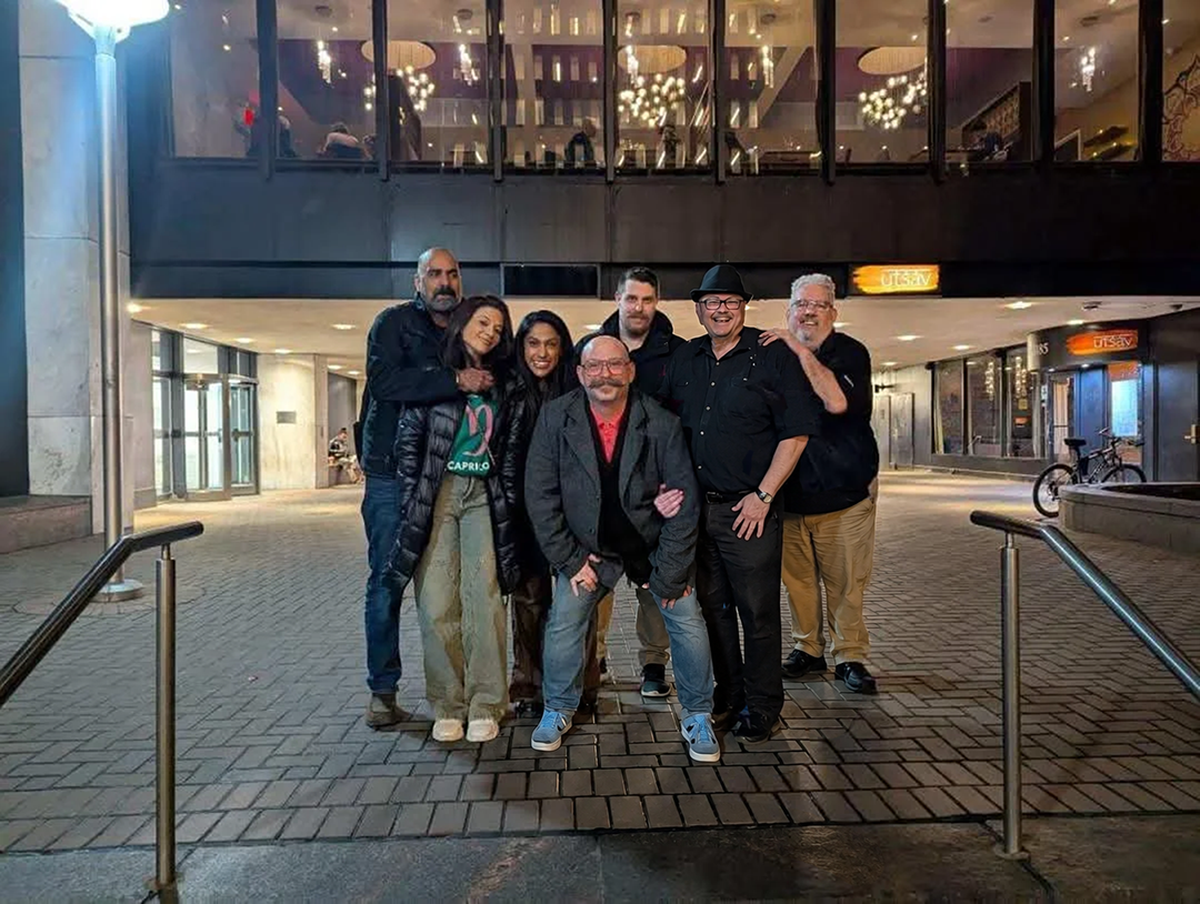 A group of seven people posing together outside a modern building at night, some smiling and some with neutral expressions. They are standing on a paved area, with large glass windows and doors visible behind them, and a staircase to the right.