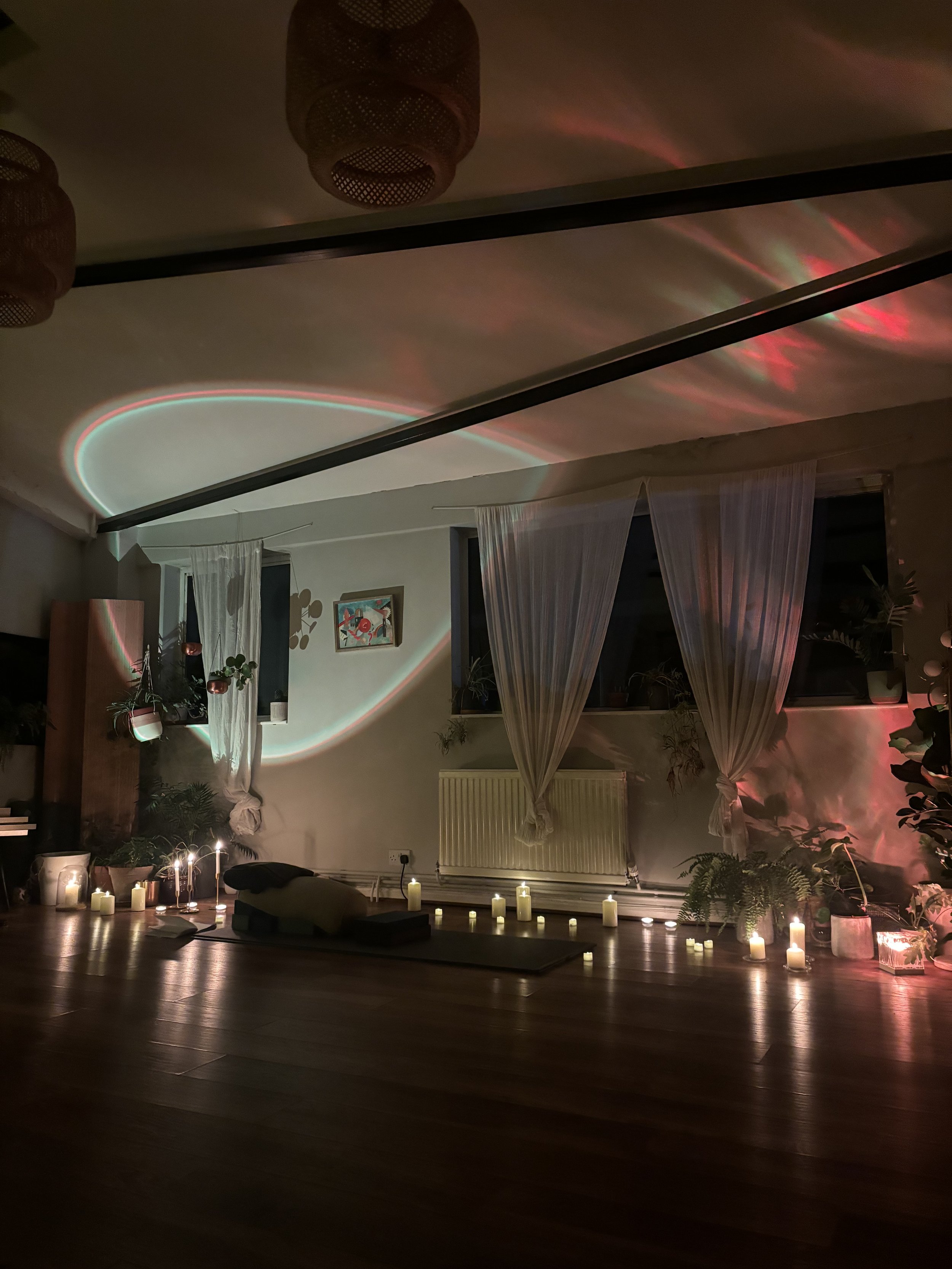 A cozy yoga or meditation space decorated with lit candles, hanging plants, sheer curtains, and ambient lighting creating a calming atmosphere.