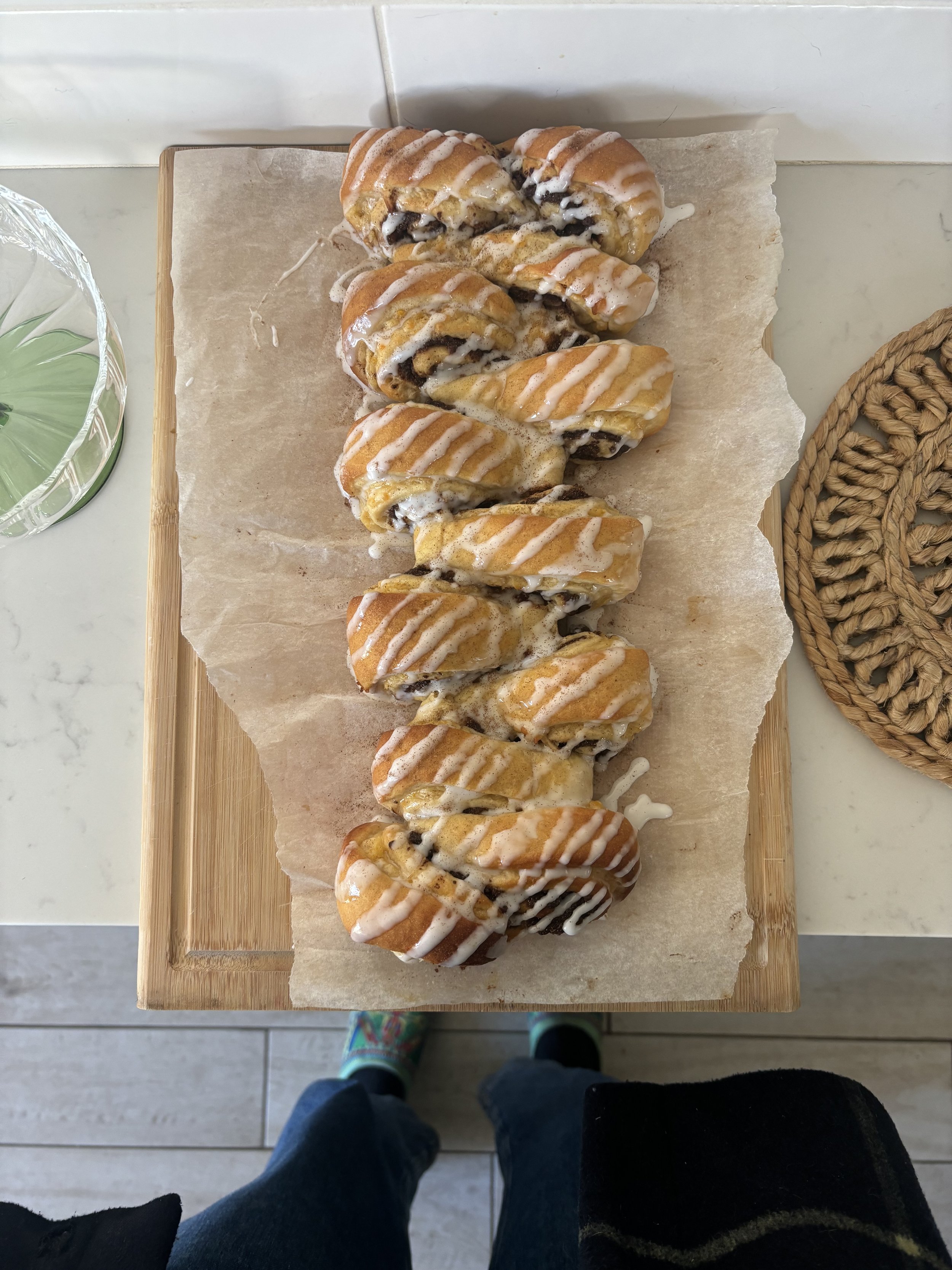 Ten cinnamon rolls with icing drizzled on top placed on parchment paper on a wooden cutting board.