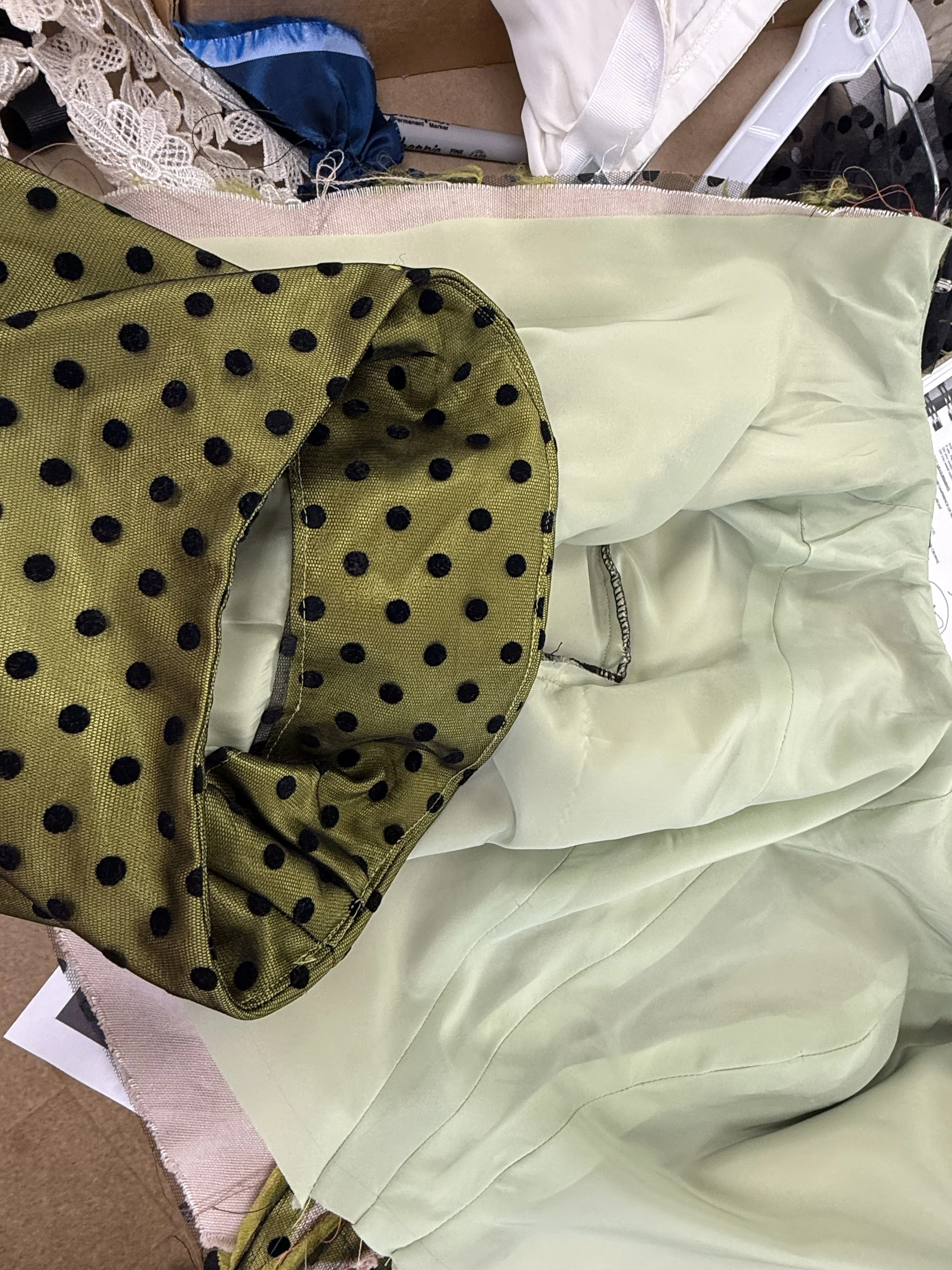 A partially constructed dress with a cream/yellowish satin fabric and a green satin collar with black polka dots, lying on a pink sewing machine cover.