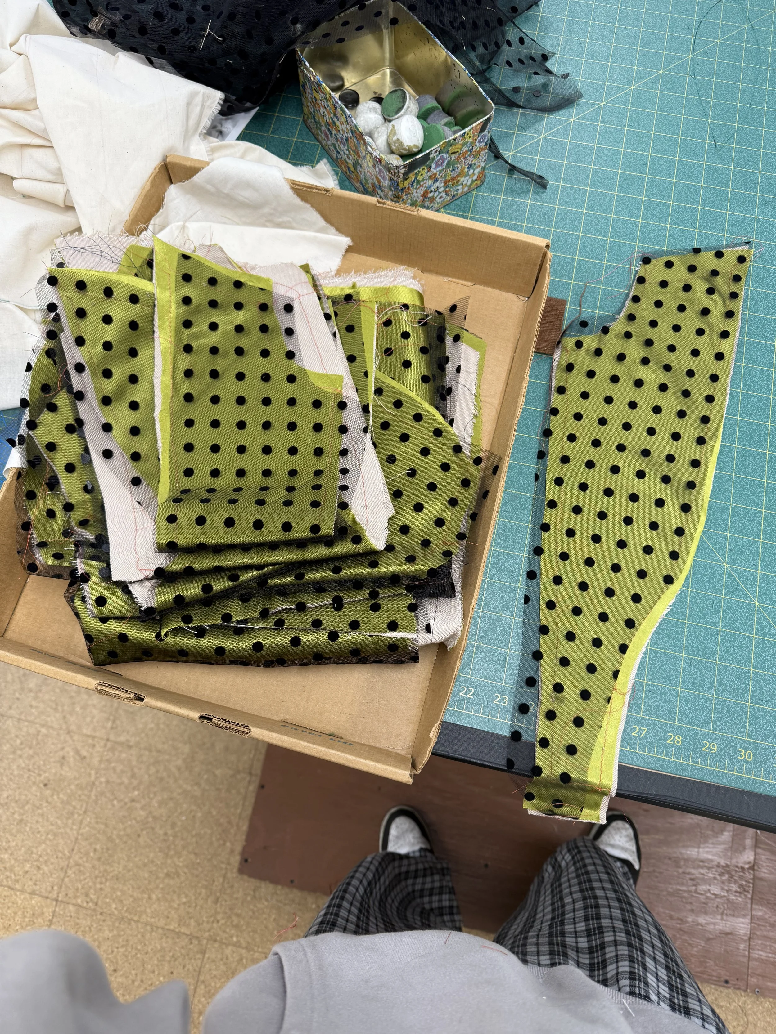 A workspace with a cutting mat, fabrics, and sewing supplies. Green polka dot fabric is cut and stacked, with some strips cut and laid out. There is a box of assorted pins, and a decorative box of buttons, with an individual wearing plaid shorts and a gray top visible at the bottom of the image.