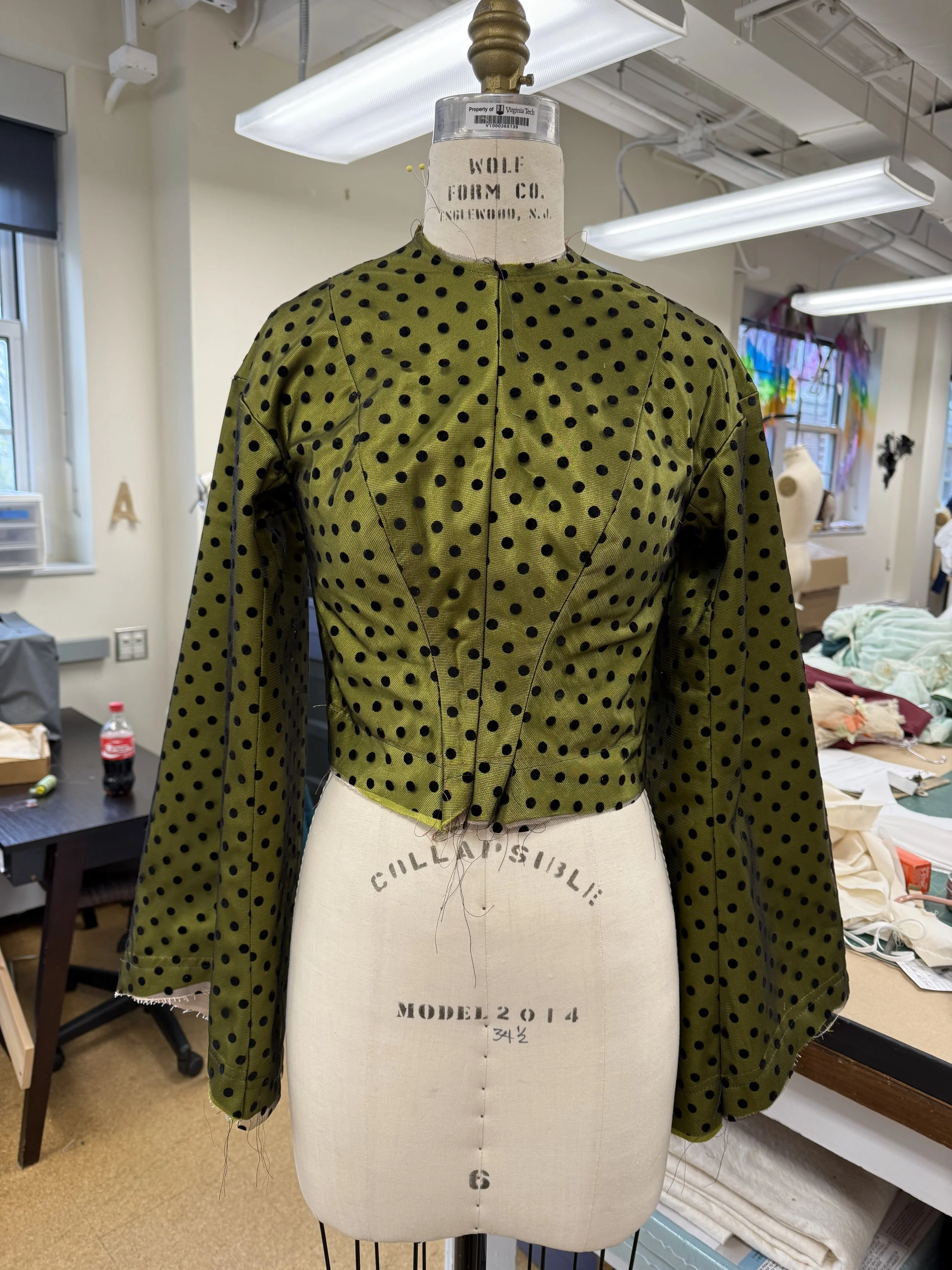 A vintage dress form mannequin wearing an olive green blouse with black polka dots, displayed in a sewing room. The dress form has labels indicating it is a 'WOLF FORM CO.' model from 2014.