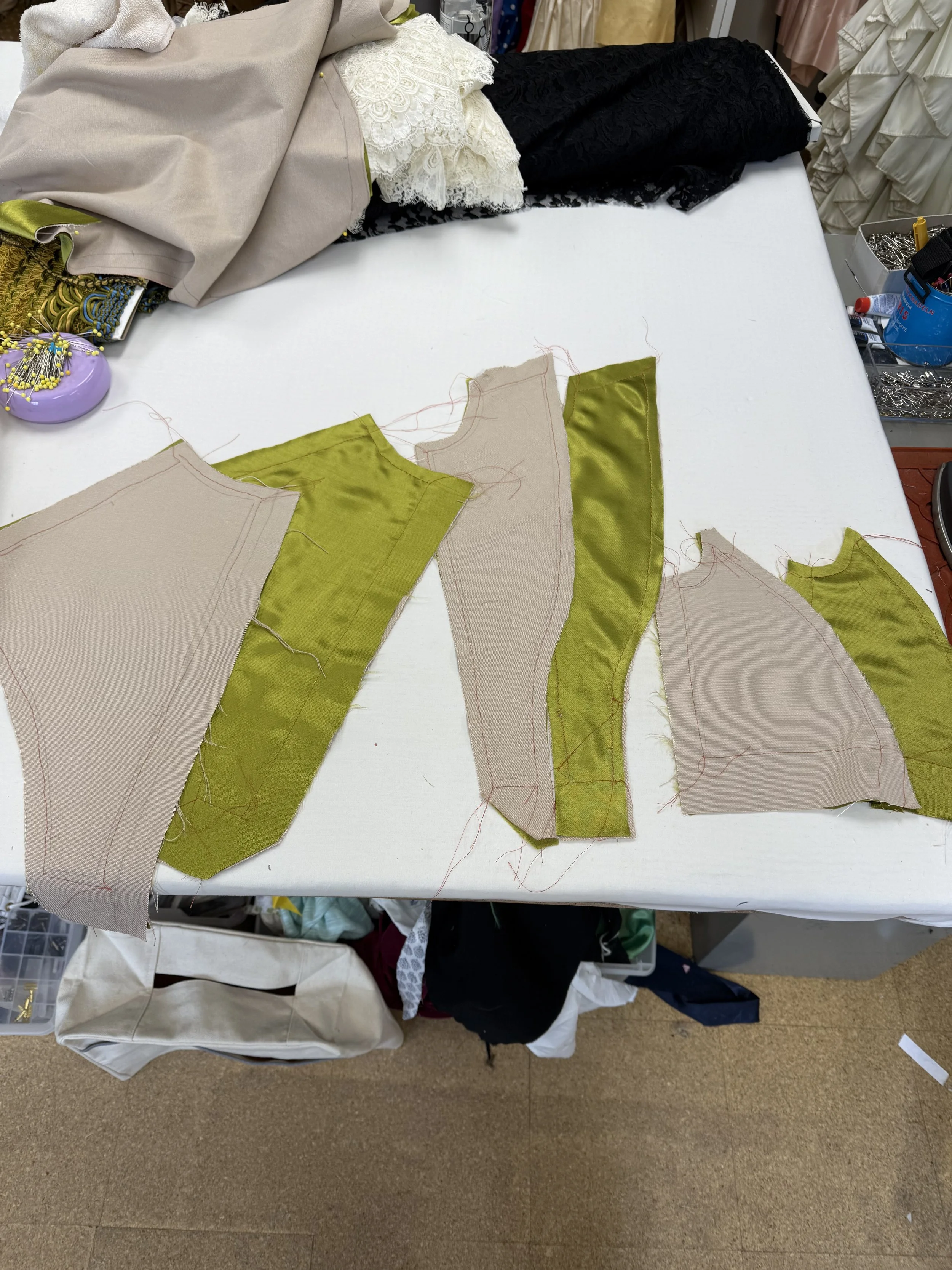 Fabric pieces cut for garment sewing, on a white worktable, mostly in beige and green colors, surrounded by sewing tools and other fabric samples.