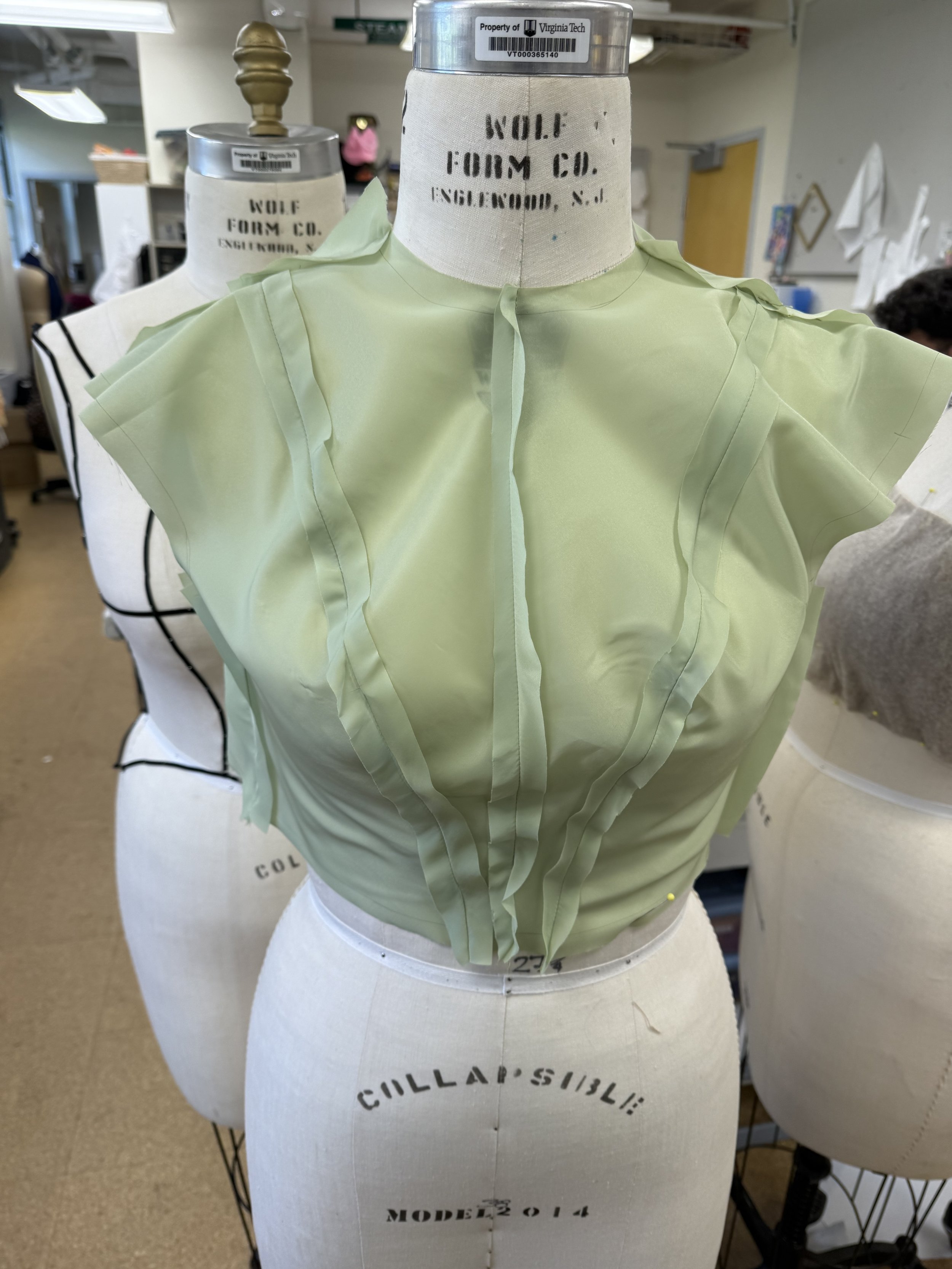 Light green sheer fabric draped on a dress form in a sewing workshop, with multiple layers and ruffles, labeled 'Wolf Form Co. Englewood, N.J.'