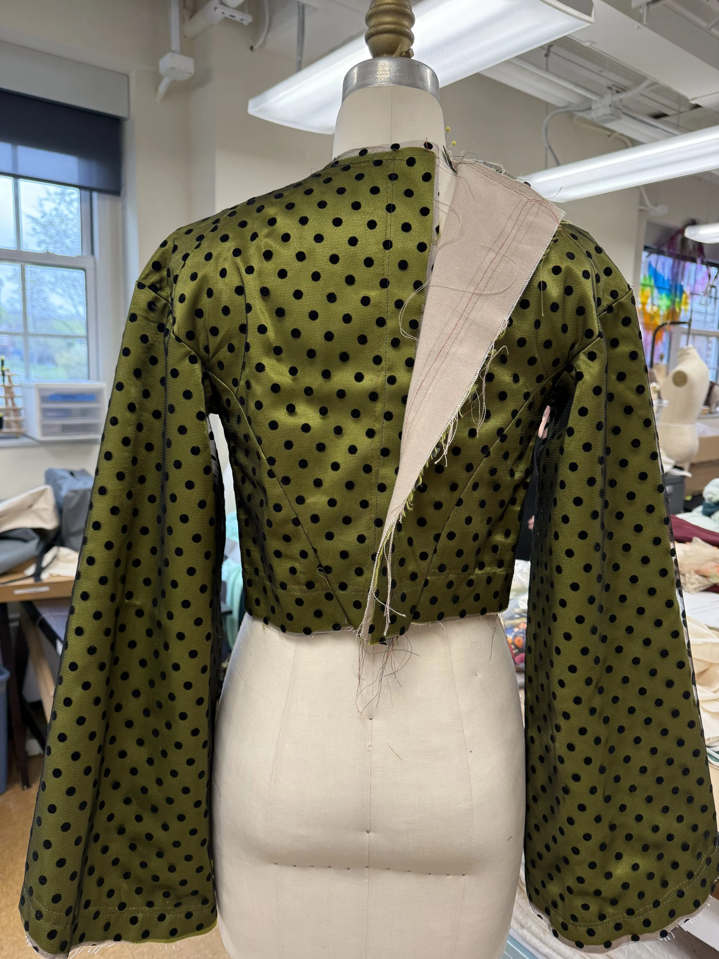 A dress form wearing an olive green, polka-dot long sleeve jacket in a sewing studio.