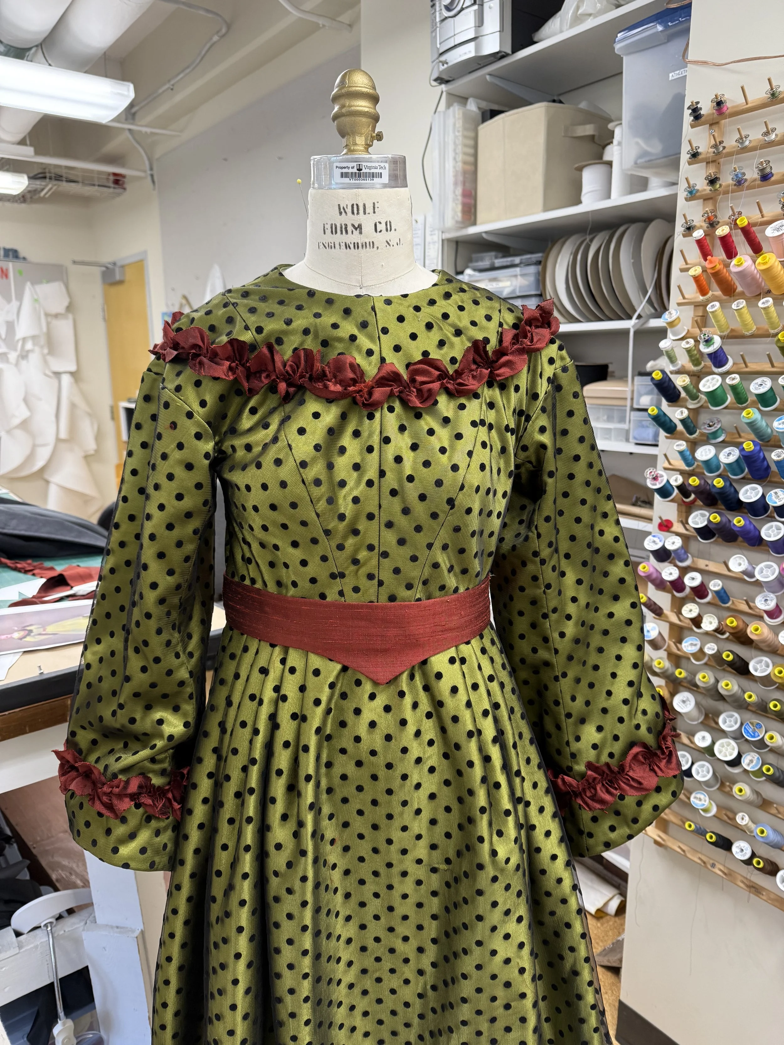A vintage green polka dot dress with red ruffle trim and a red waistband displayed on a dress form in a sewing room.