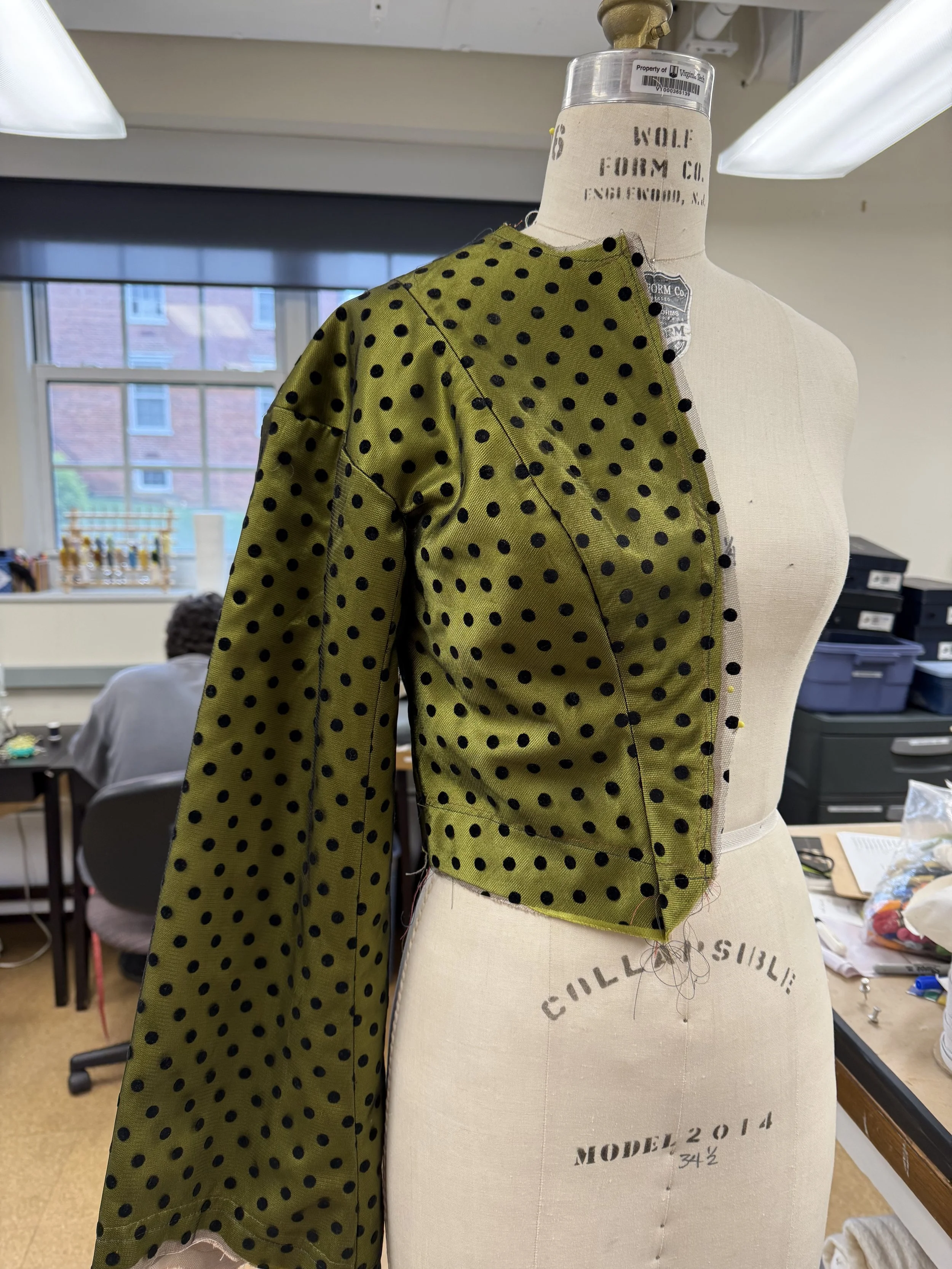 Partial view of an olive green fabric with black polka dots draped over a dress form or mannequin, with colorful workspace and person in the background.