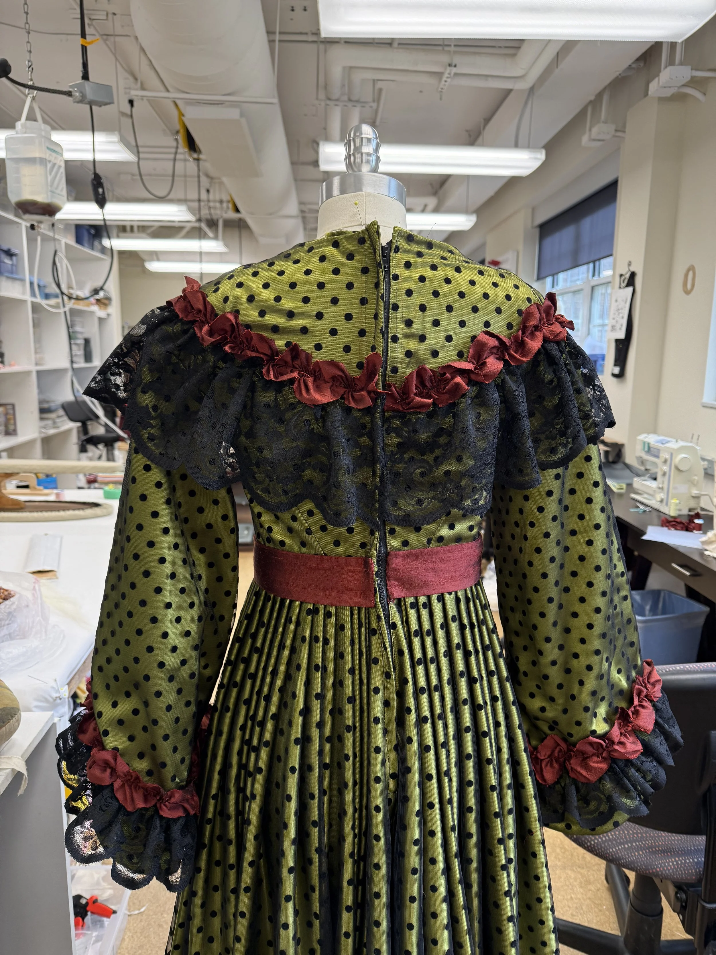 A vintage-style dress on a mannequin with a green fabric featuring black polka dots, black lace details on the shoulders and cuffs, red ruffles around the neckline and waist, in a room with sewing and craft supplies.
