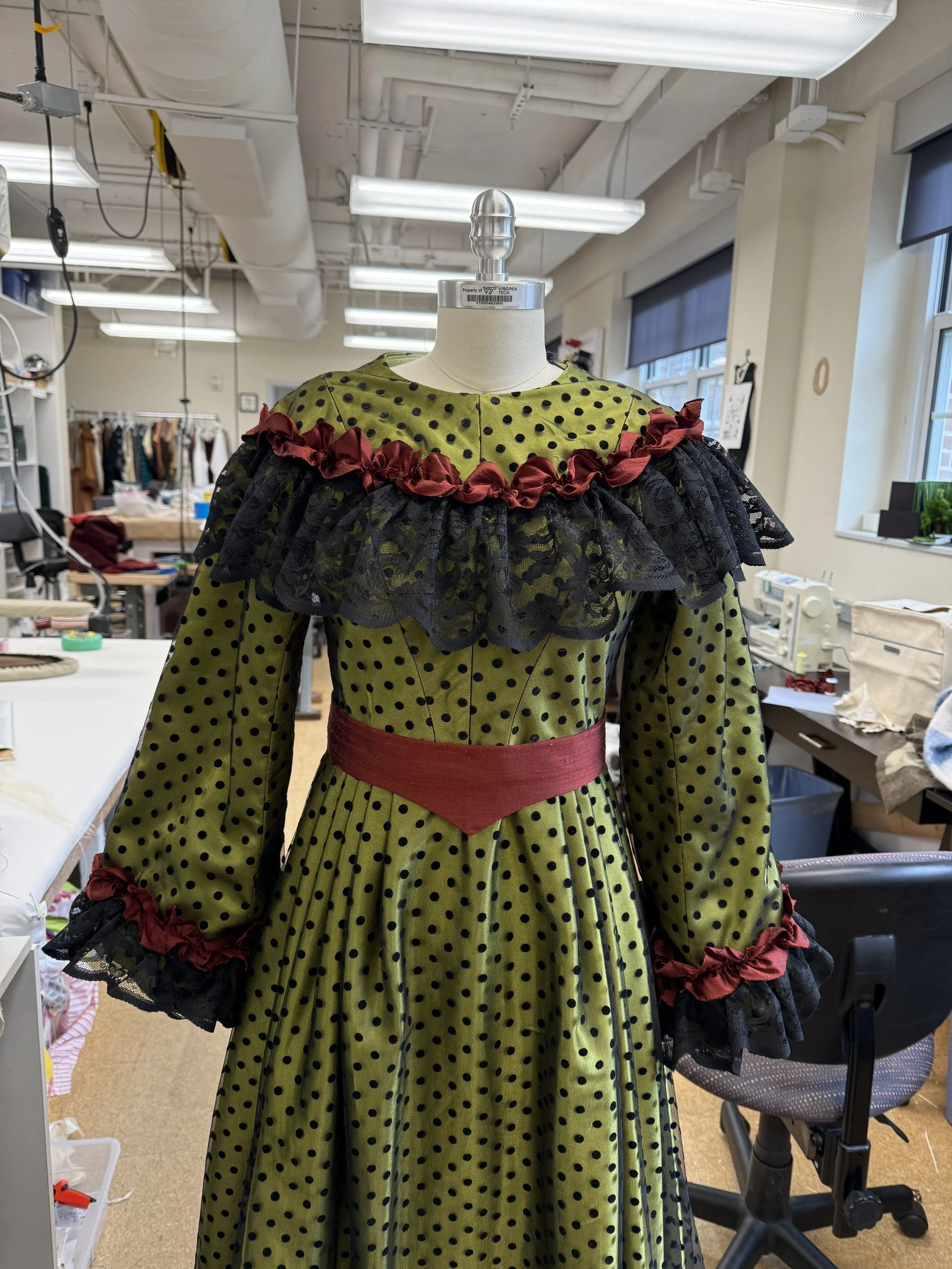 A mannequin wearing a vintage-style, green dress with black polka dots, black lace and red accents. The dress features a ruffled black lace collar with red trim, long sleeves with ruffled black lace cuffs, and a red waistband. The mannequin is in a sewing or costume workshop with fabric, sewing tools, and furniture in the background.