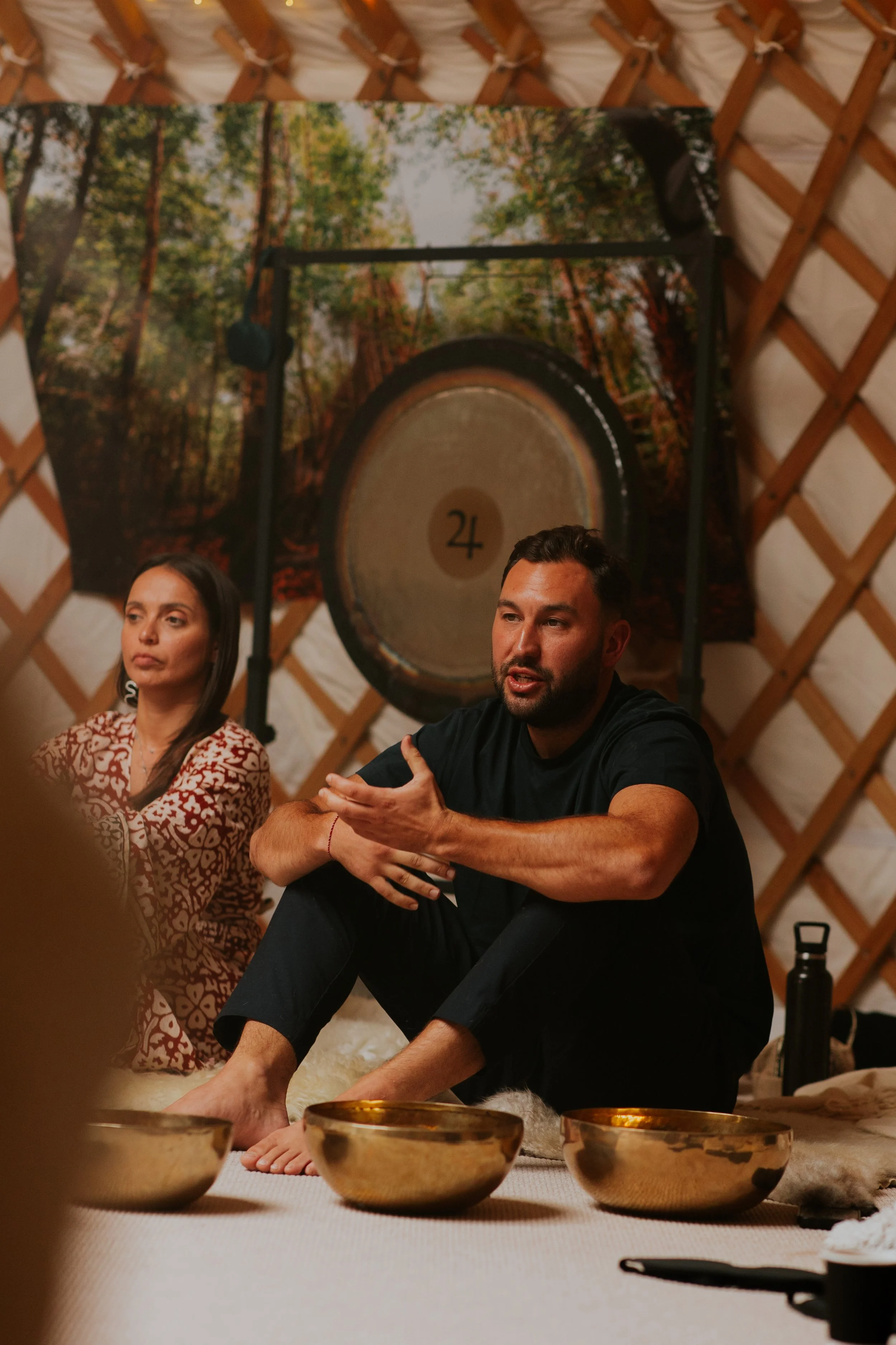A man speaking with hand gestures sitting on the floor next to a woman listening. There are singing bowls in front of him and a large drum hanging on the wall behind them, which features a forest scene.