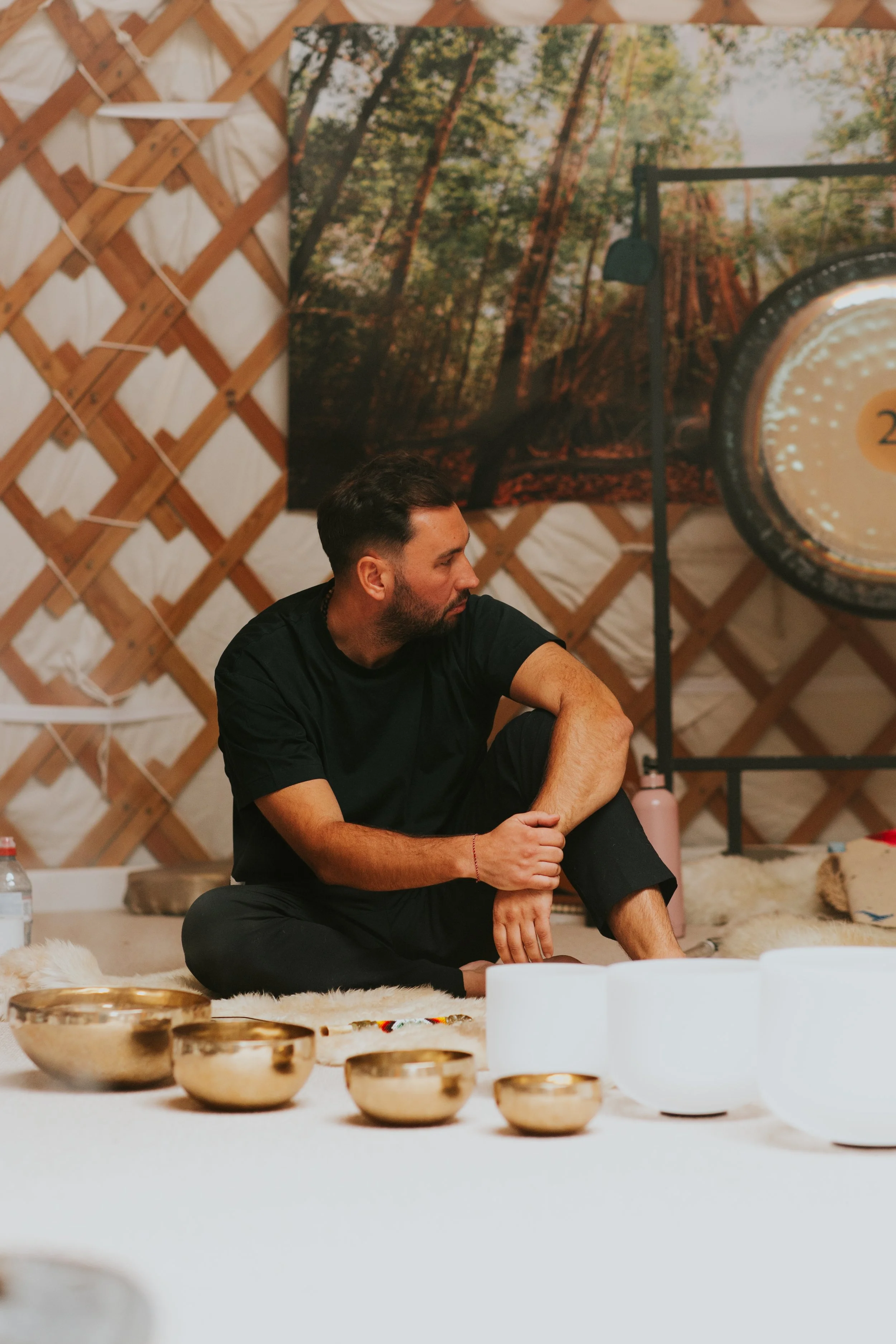 A man sitting on the floor among singing bowls and candles inside a yurt, with a forest scene artwork on the wall behind him.