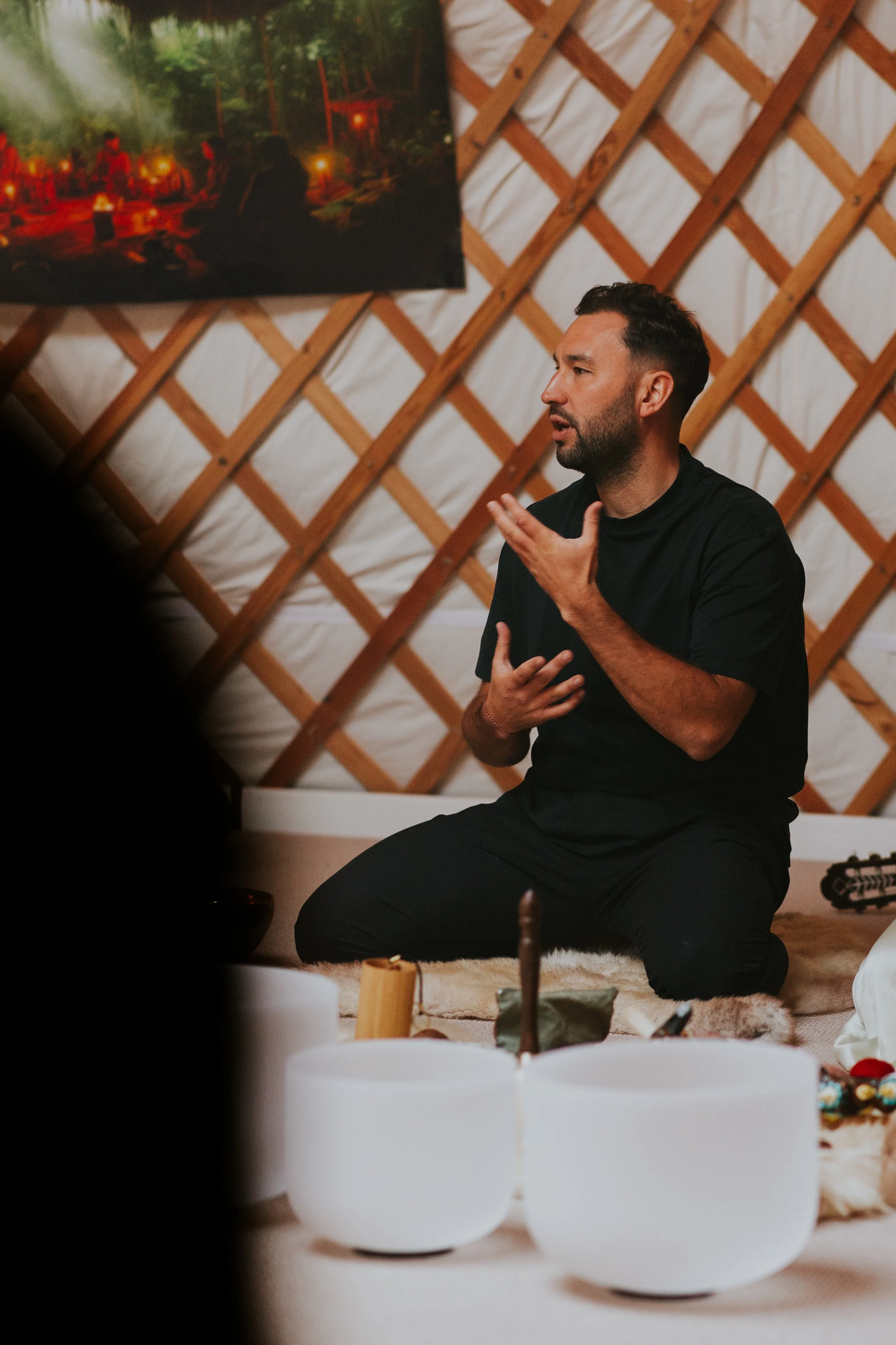 A man kneeling on the floor, wearing a black shirt, speaking or singing passionately with one hand on his chest and the other near his face, in a yurt with wooden lattice walls. There are singing bowls in front of him and a painting of a campsite scene hanging on the wall behind.