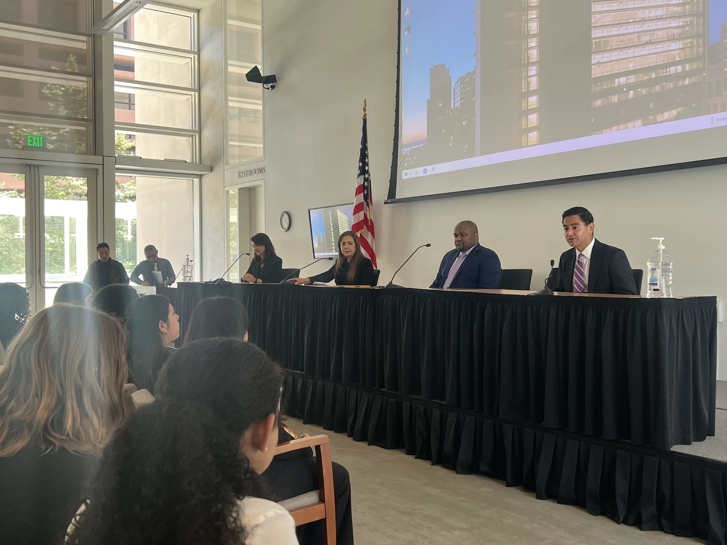 A panel of five people seated at a long black table on a stage during a conference or seminar. There is an American flag behind the panel, and a large screen displaying cityscape images above them. Audience members are seated and listening attentively.