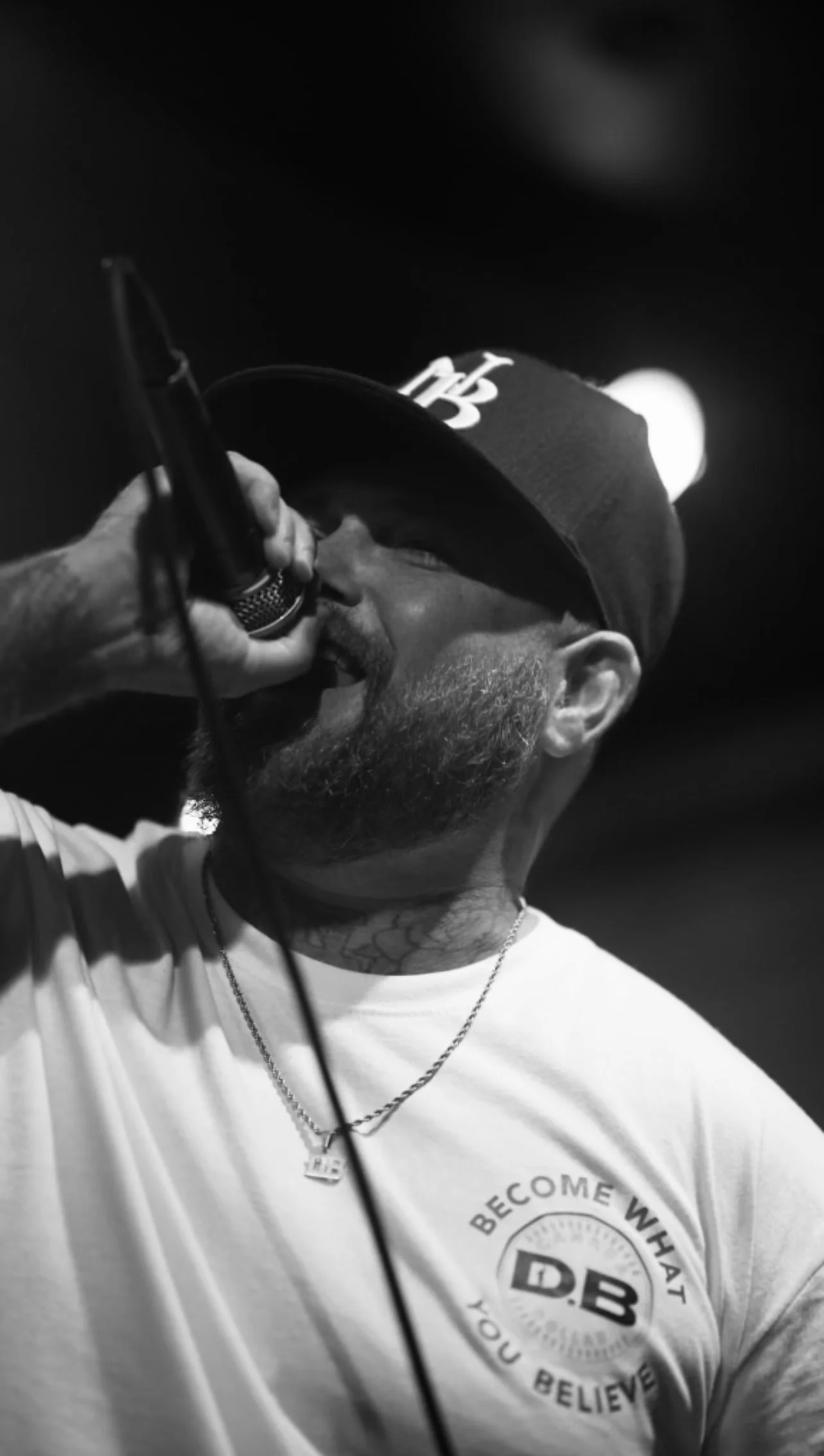 A man with a beard and tattoo on neck, wearing a cap with 'DB' logo, a white t-shirt with matching logo, and a chain necklace, singing or speaking into a microphone on stage.