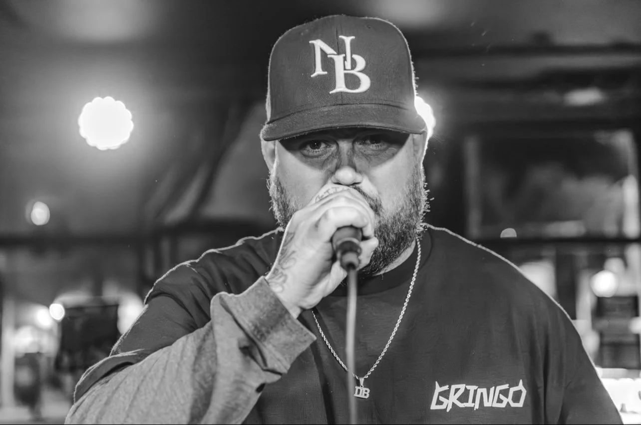 A man singing into a microphone on stage, wearing a baseball cap and a shirt with 'Gringo' on it, in black and white.