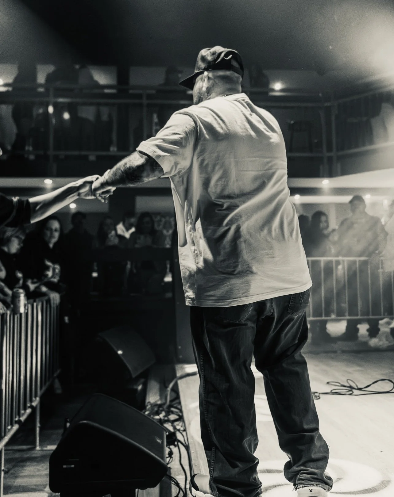 A performer in casual clothing and a cap interacts with the audience on stage during a live music event, with a crowd watching in the background.