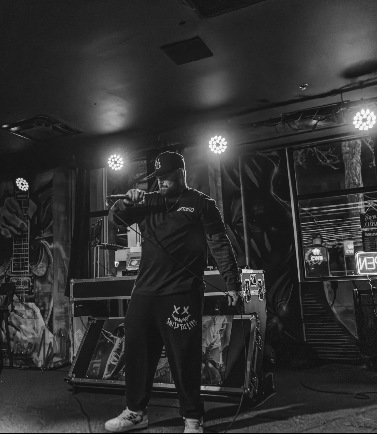 Male performer in hoodie and cap holding microphone on stage with DJ equipment, bright lights above, graffiti on walls behind him, in black and white.