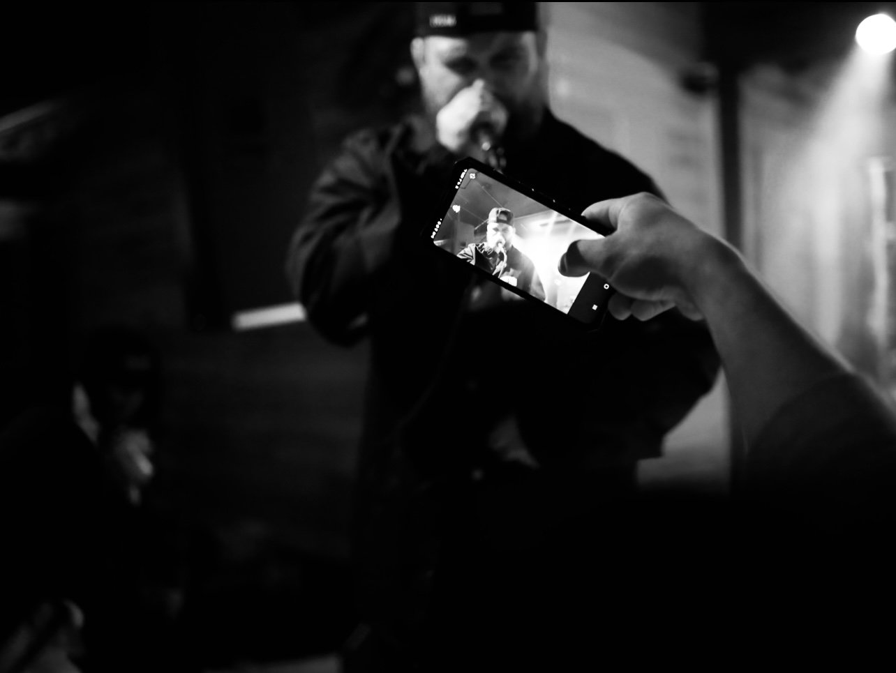 A person taking a photo of a male singer with a smartphone during a performance, in black and white.