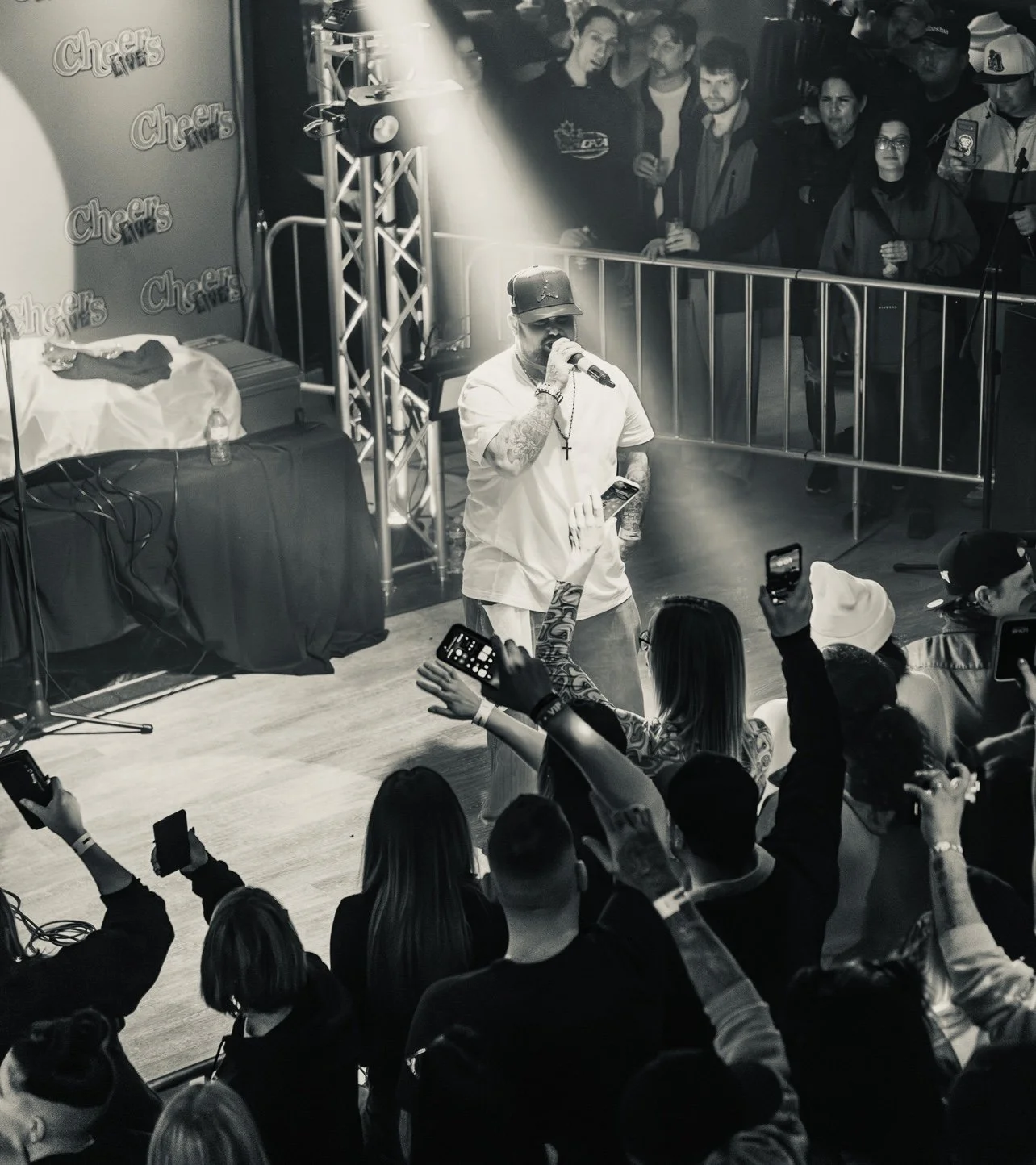 A male performer with tattoos, wearing a cap, white T-shirt, and cross necklace, singing into a microphone on stage at a concert or event, with an audience of people taking photos and videos.