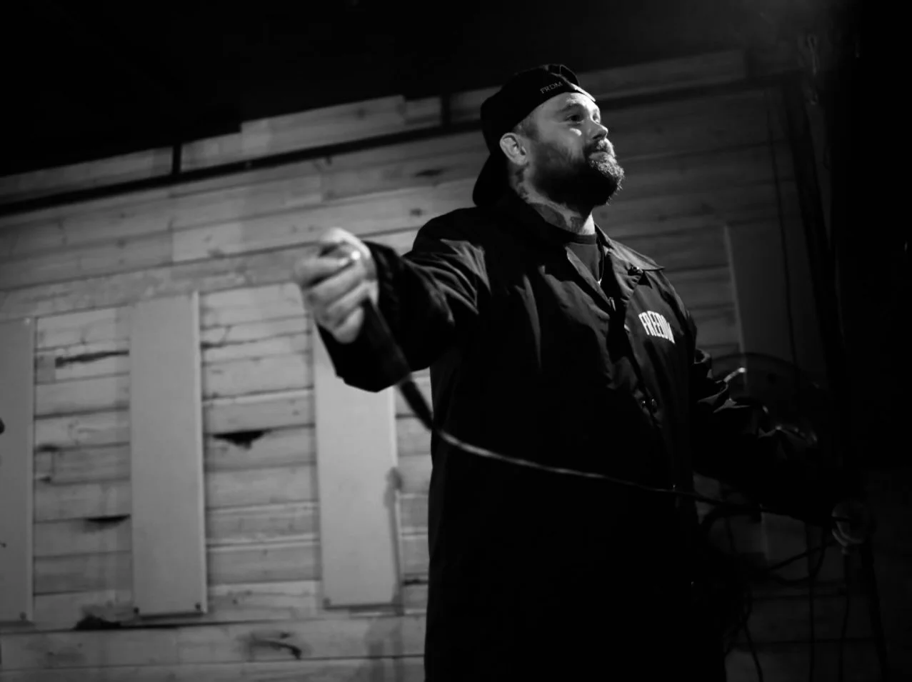 Black and white photo of a man with a beard and tattoos, wearing a backwards cap and a jacket with 'FREEDOM' on it, gesturing or speaking in an indoor setting with wooden wall panels.