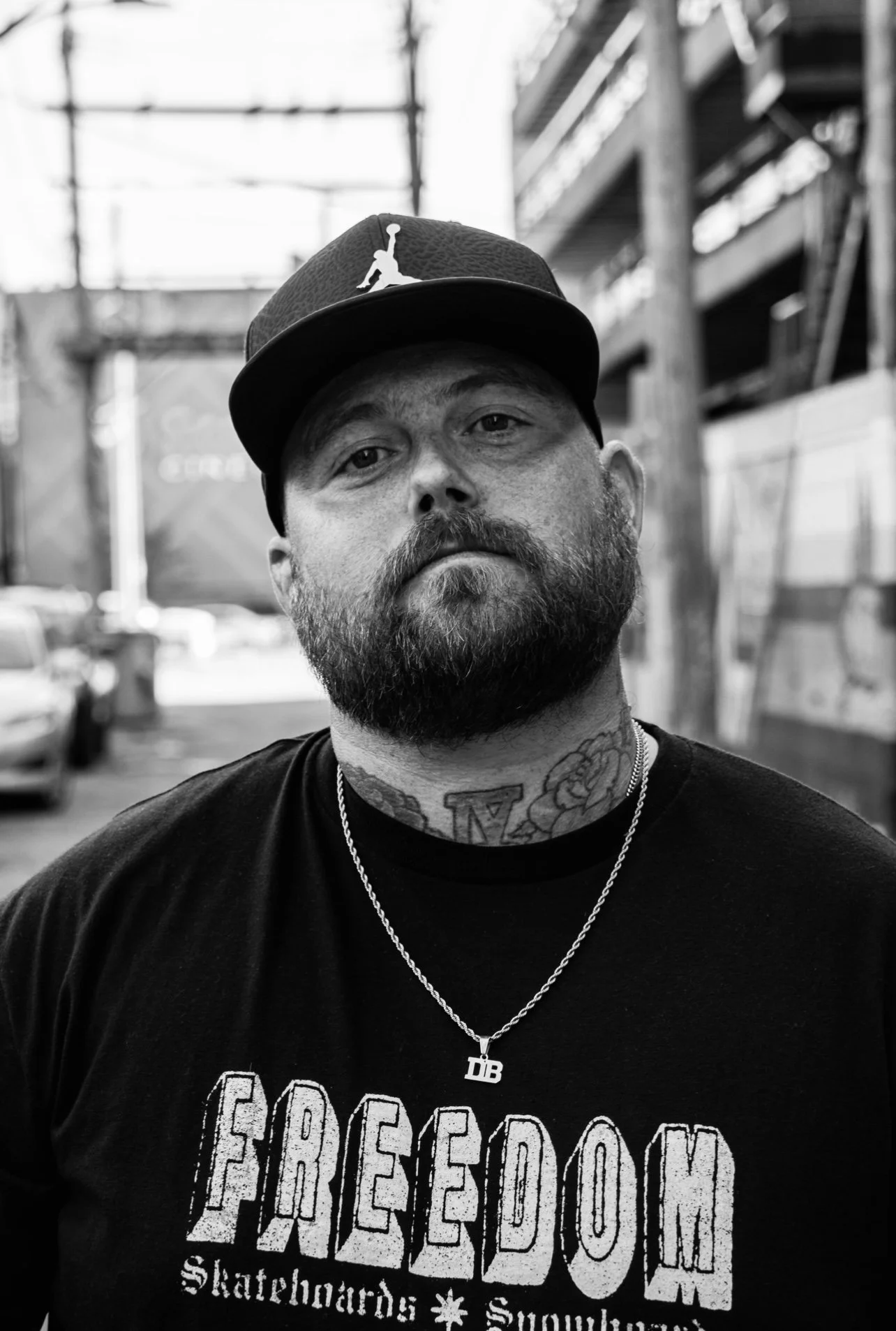A man with a beard and tattoos wearing a baseball cap and a black T-shirt that reads "FREEDOM Skateboards". He has a chain necklace with a pendant that reads "DB". The background shows an outdoor urban setting with a bridge and buildings.