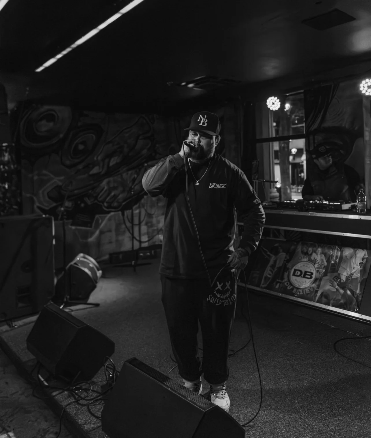 A black-and-white photo of a man singing into a microphone on stage in a music venue, with a DJ in the background, illuminated by stage lights.