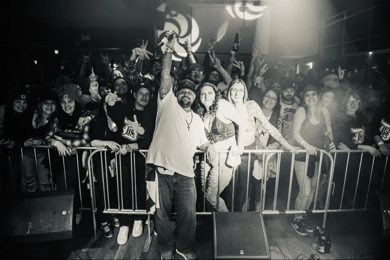 Crowd at concert or event, with people smiling and making hand signs, electronic equipment in front, and a man in the foreground holding a microphone.