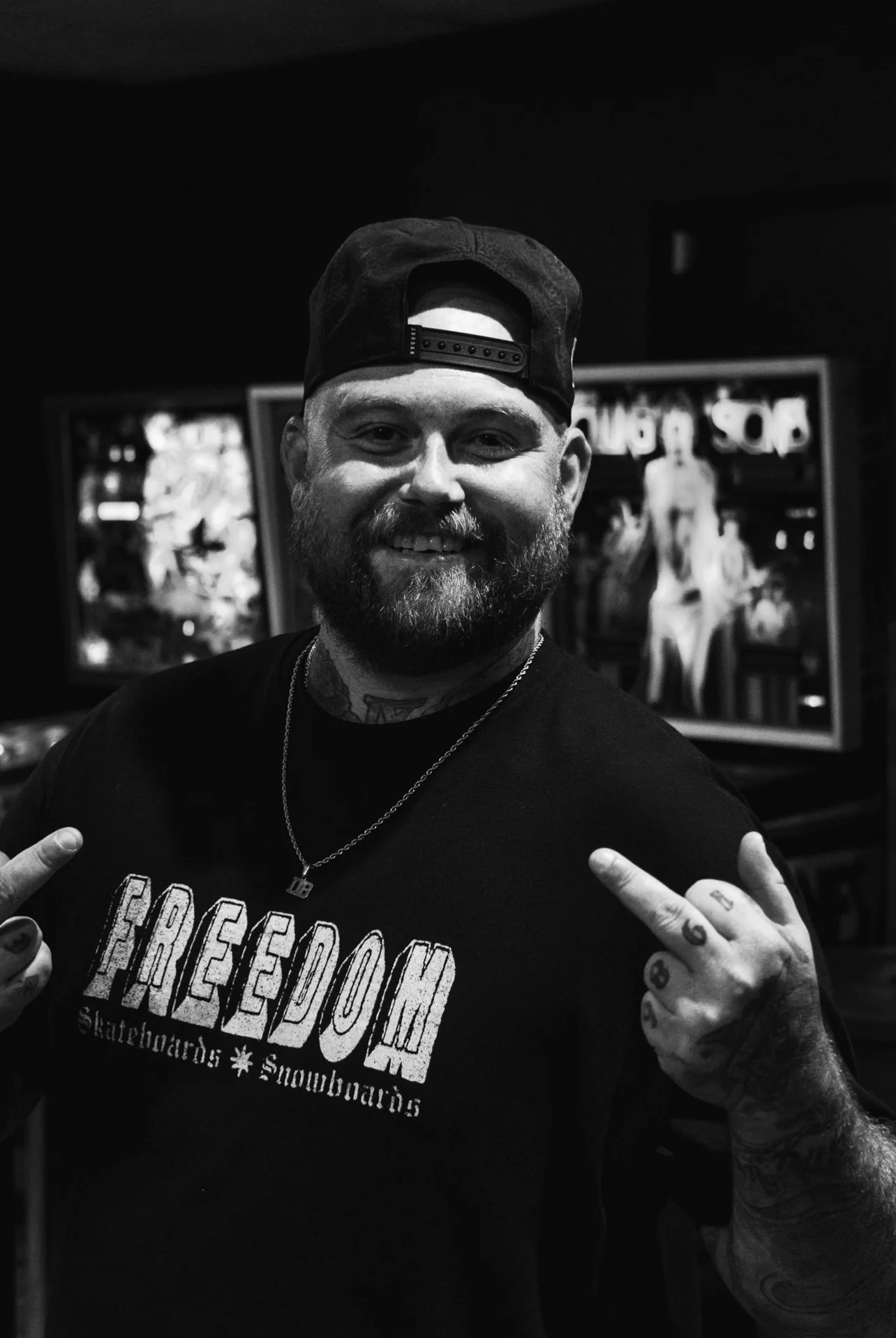 A smiling man with a beard, wearing a cap backwards, showing tattoos on his hands and neck, and pointing to his shirt that reads 'FREEDOM', with video game pinball machines in the background.