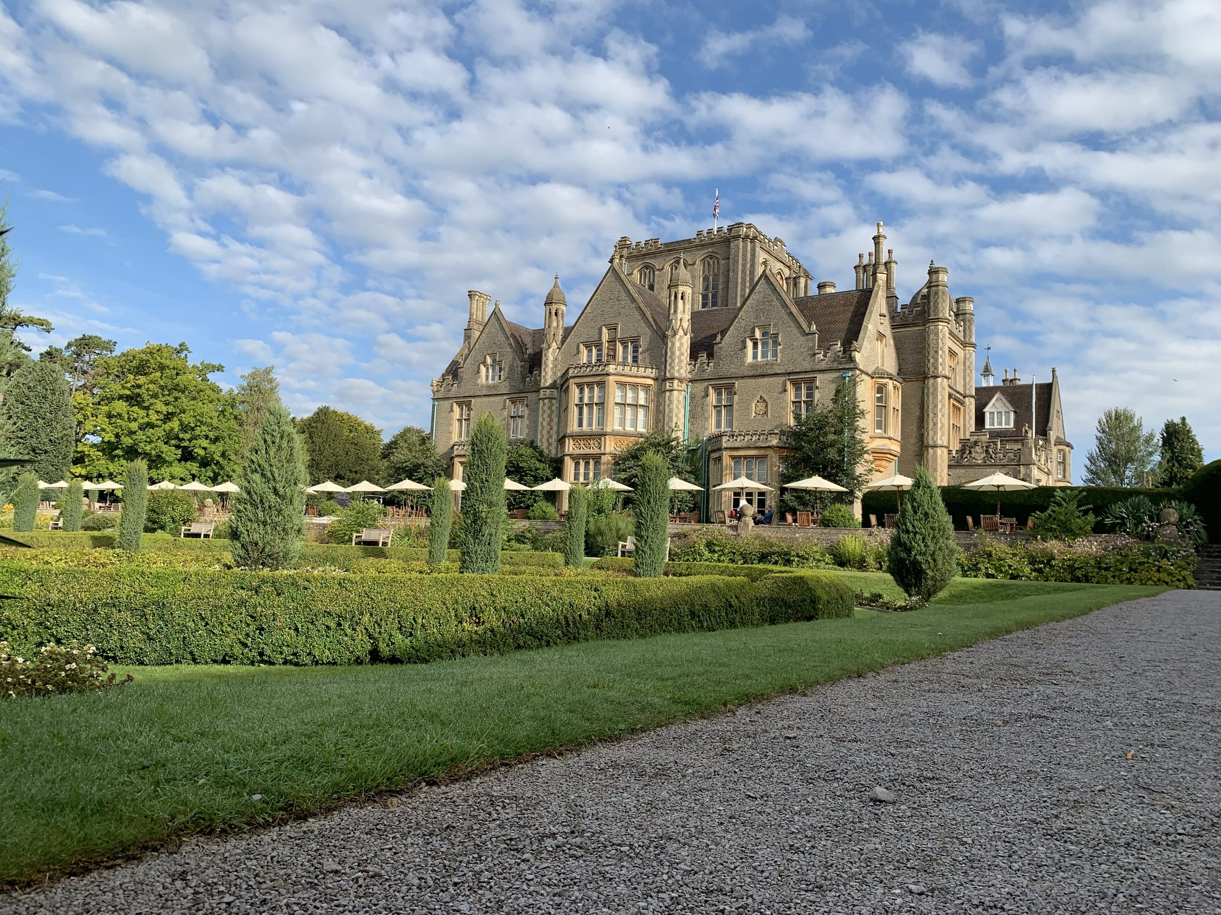 A large historic castle with pointed towers and a stone facade, surrounded by a landscaped garden with trimmed hedges and trees, under a partly cloudy sky in the Cotswolds in England.