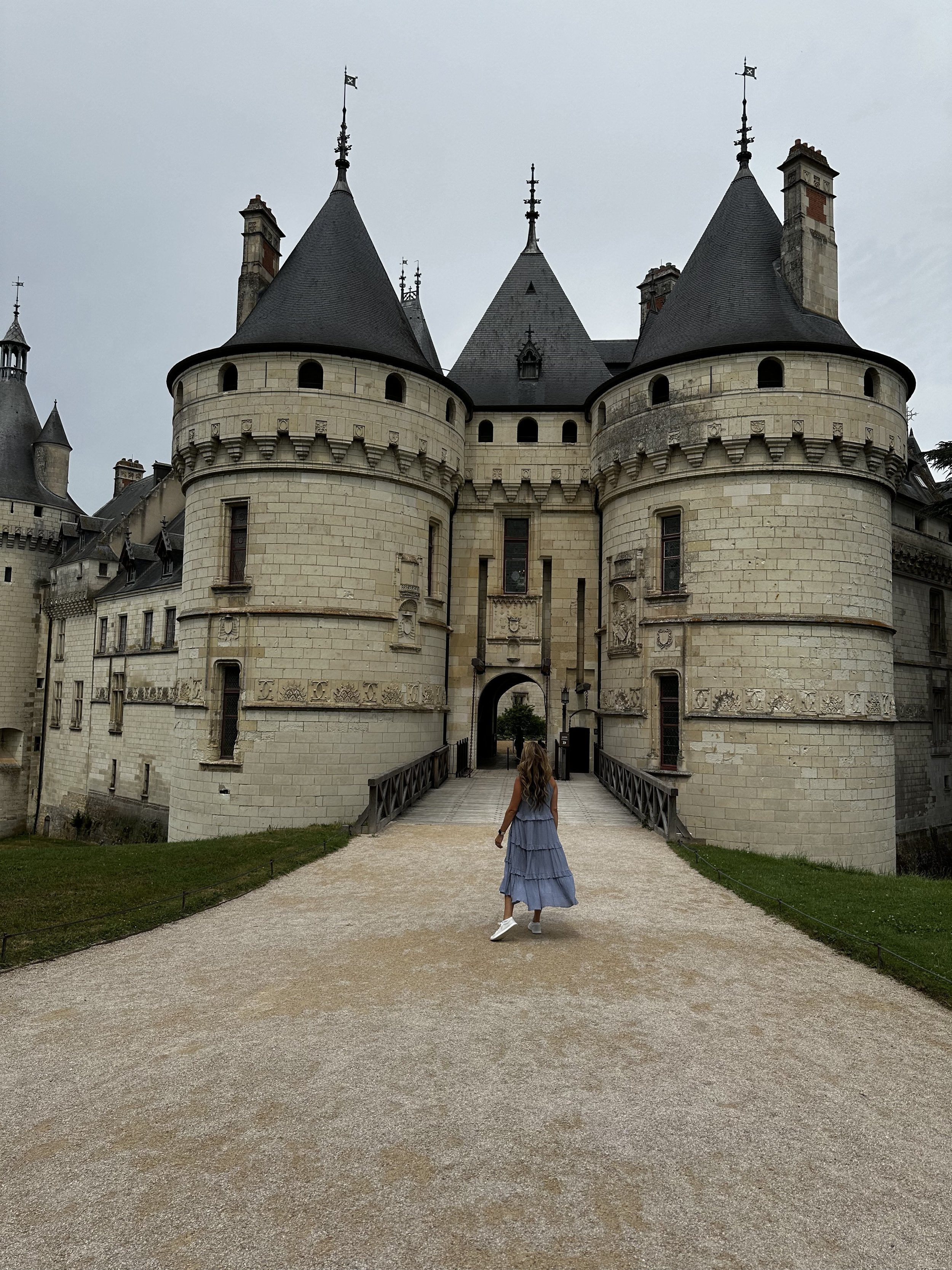 A woman in a blue dress and white shoes walking towards a castle with round towers and flags on top, over a gravel path in the Loire Valley in France.