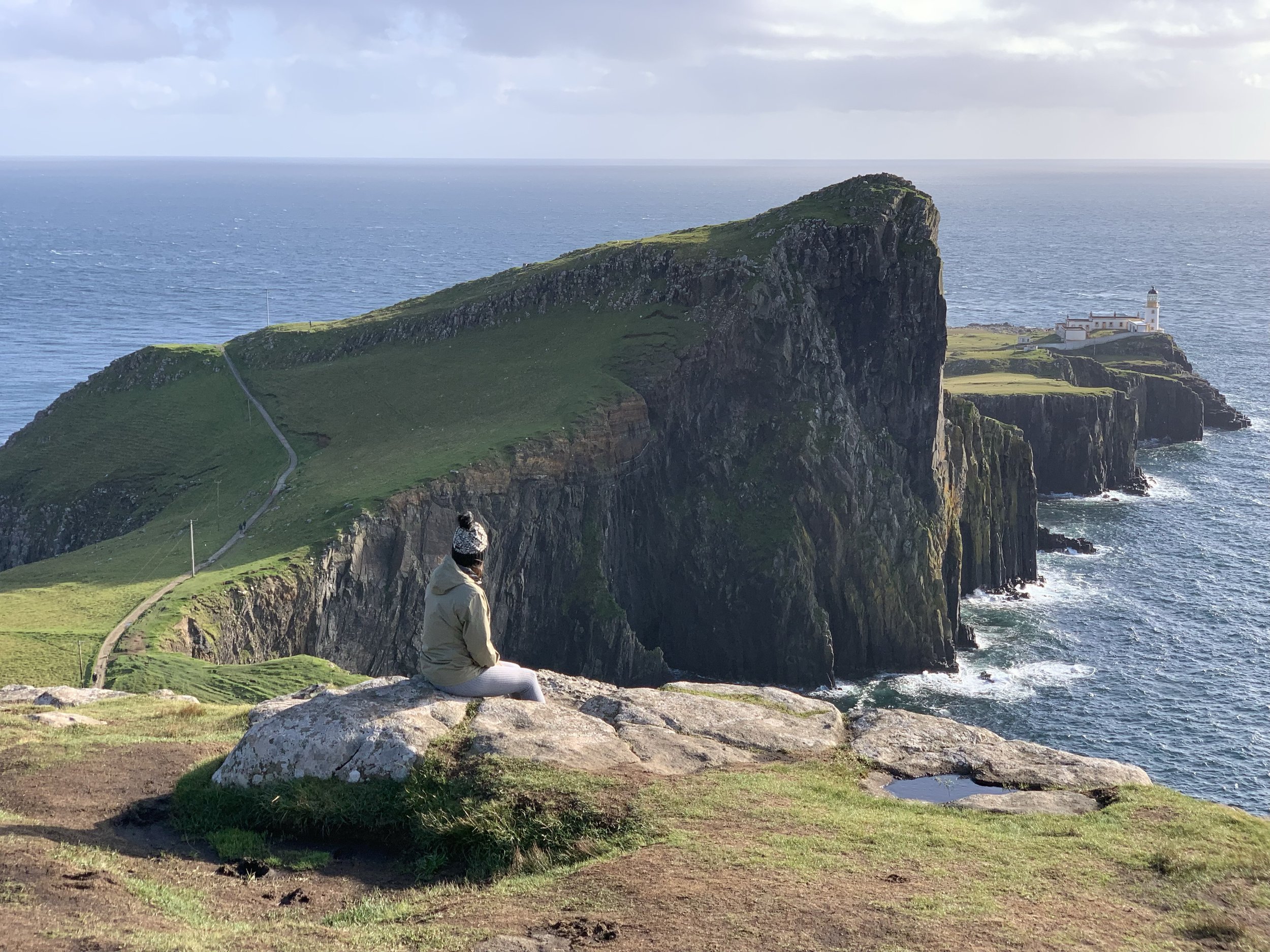A person sitting on a rock overlooking green cliffs and the ocean, with a lighthouse on a distant peninsula in Isle of Skye Scotland.