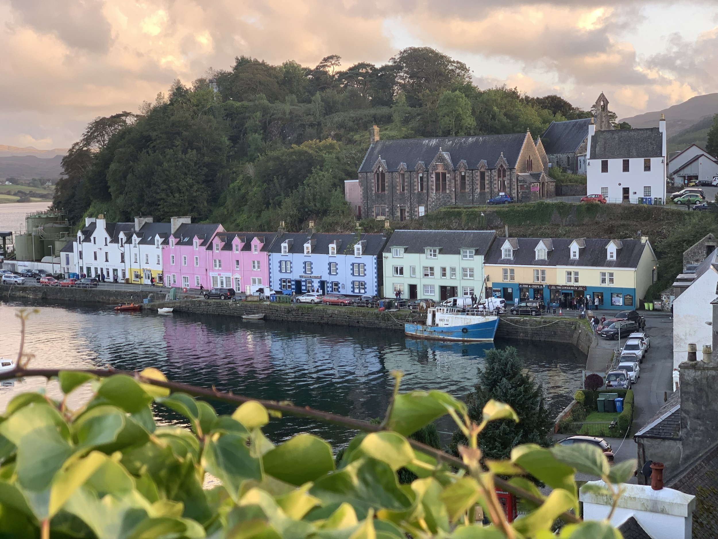 Colorful houses along a waterfront with a boat, a historic church on a hill, and lush green trees in a scenic seaside village at sunset in Portree Scotland on the Isle of Skye.