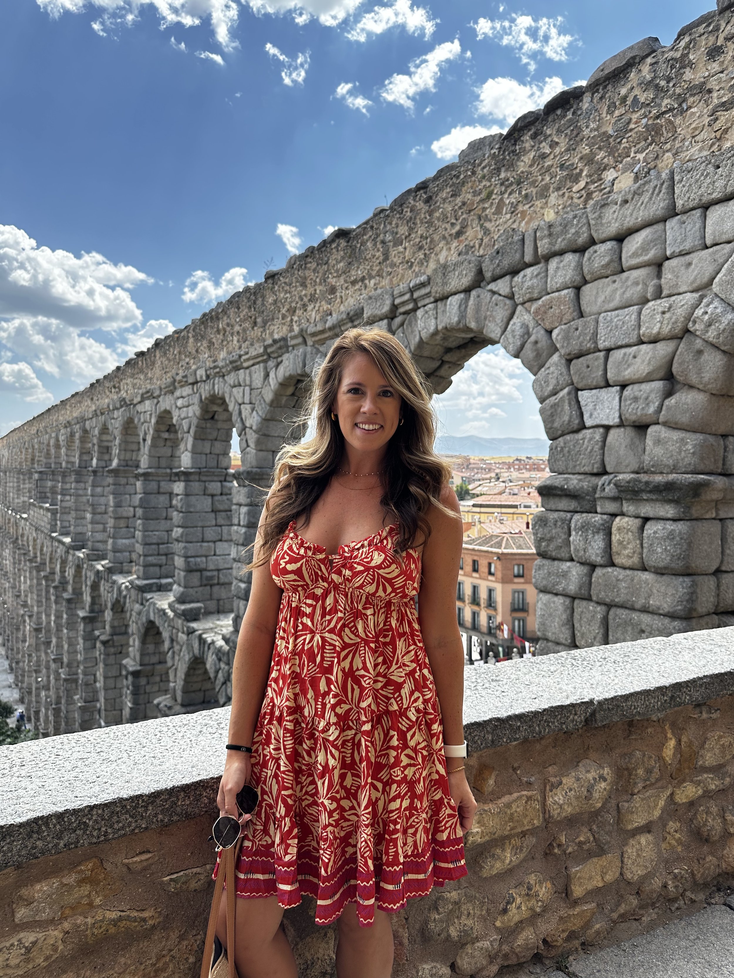 A woman in a red and beige patterned dress standing in front of the ancient Roman aqueduct in Segovia, Spain, with a bright blue sky and fluffy clouds above.