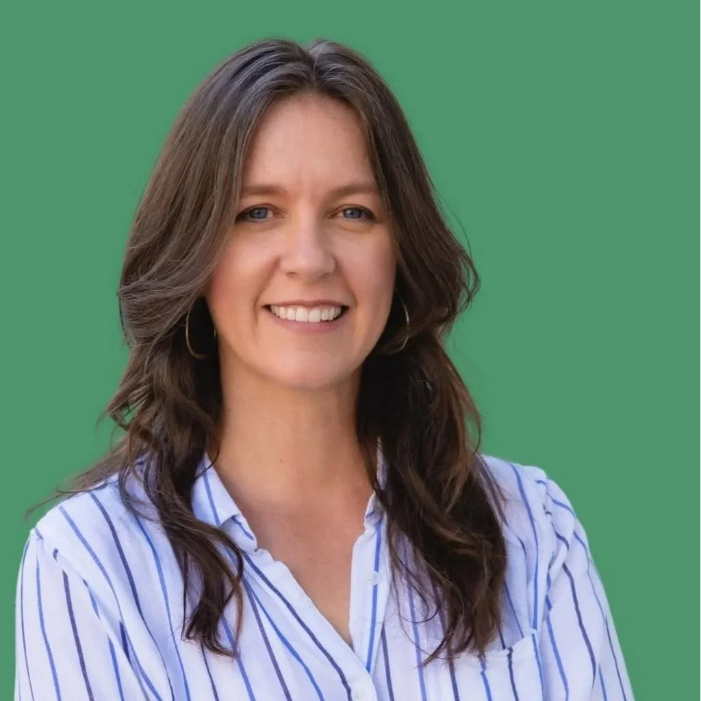 A smiling woman with wavy brown hair wearing a blue and white striped shirt, standing against a green background.