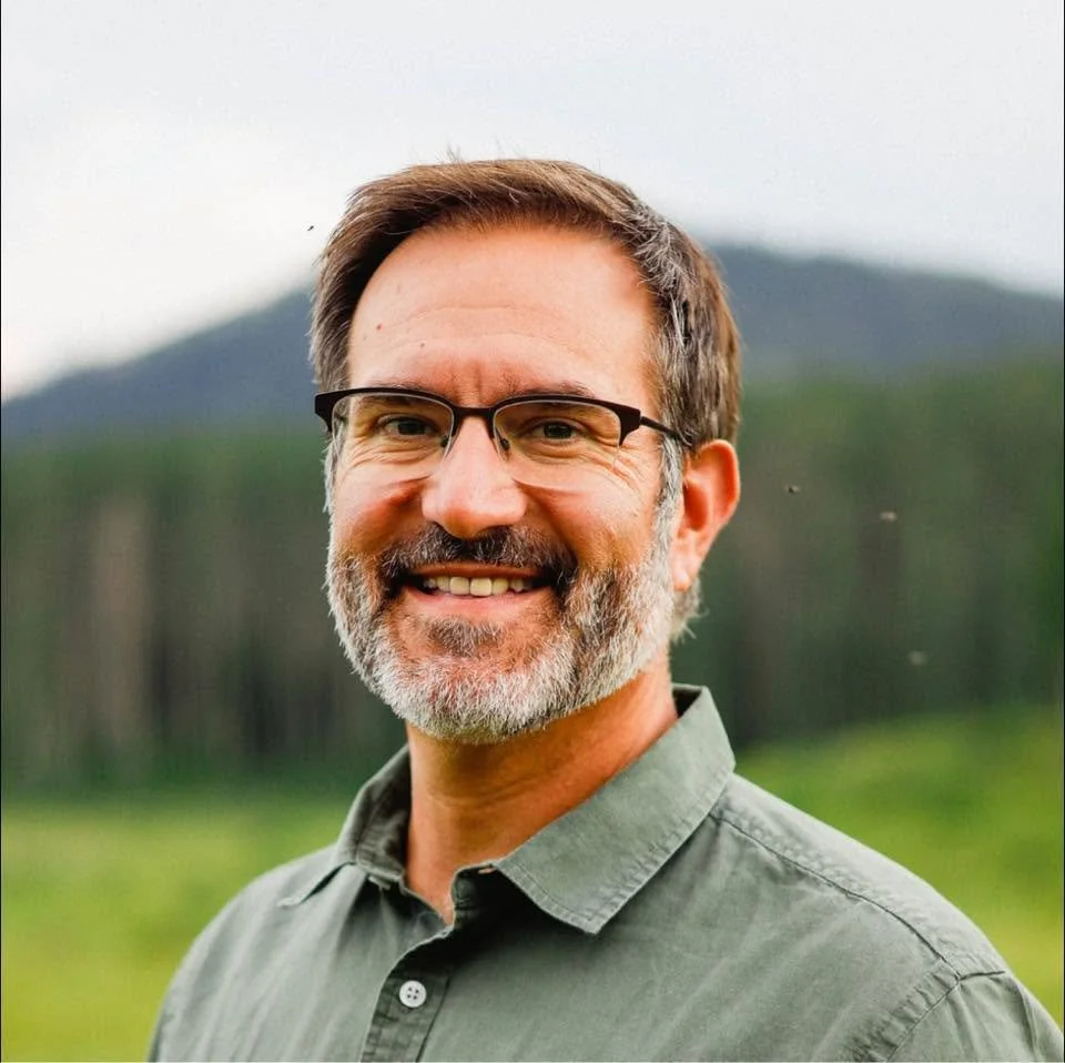 A middle-aged man with glasses, a beard, and a smile, outdoors in a green field with trees and mountains in the background.