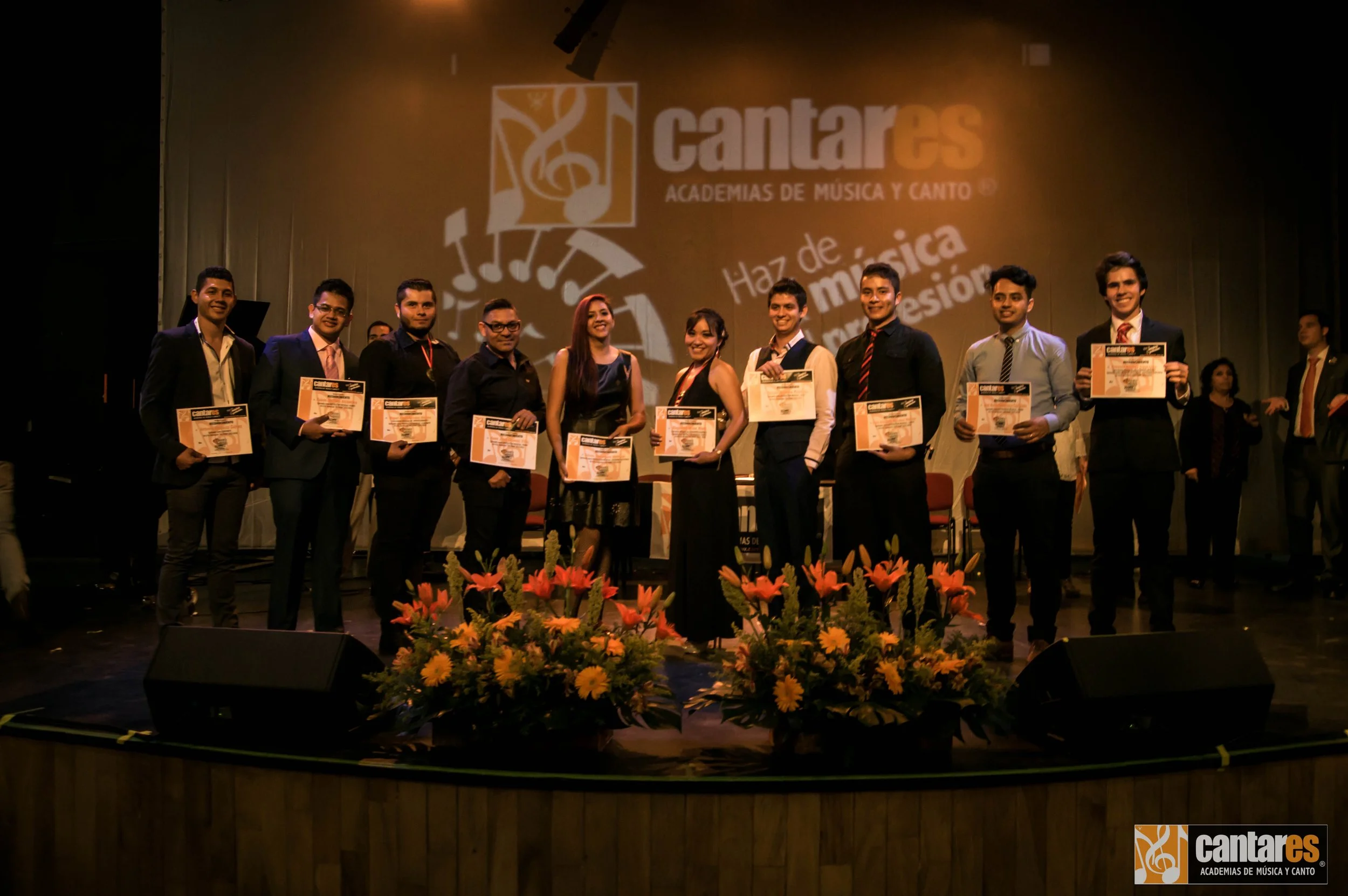 Grupo de personas en un escenario recibiendo certificados en un evento de academias de música y canto, con el logo de Cantares al fondo y arreglos florales en el frente.