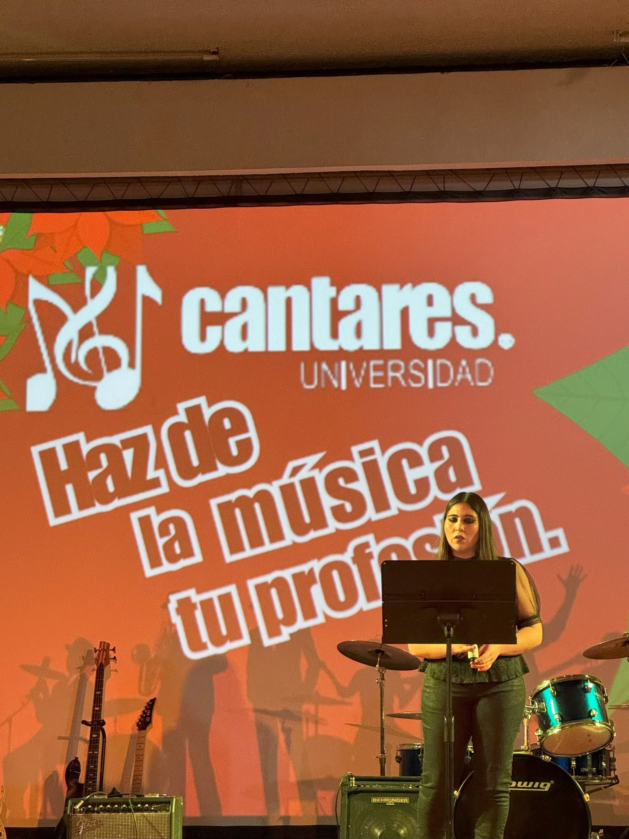 Una mujer tocando en una banda en un escenario, con un fondo que promociona un evento universitario llamado 'Cantares' y el lema 'Haz de la música tu profesión'.
