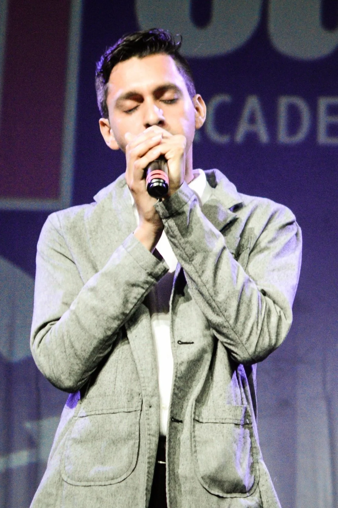 Un hombre joven con cabello oscuro y peinado, vestido con saco gris claro, canta con un micrófono en las manos cerradas, con los ojos cerrados, en un escenario con fondo azul y letras parcialmente visibles.
