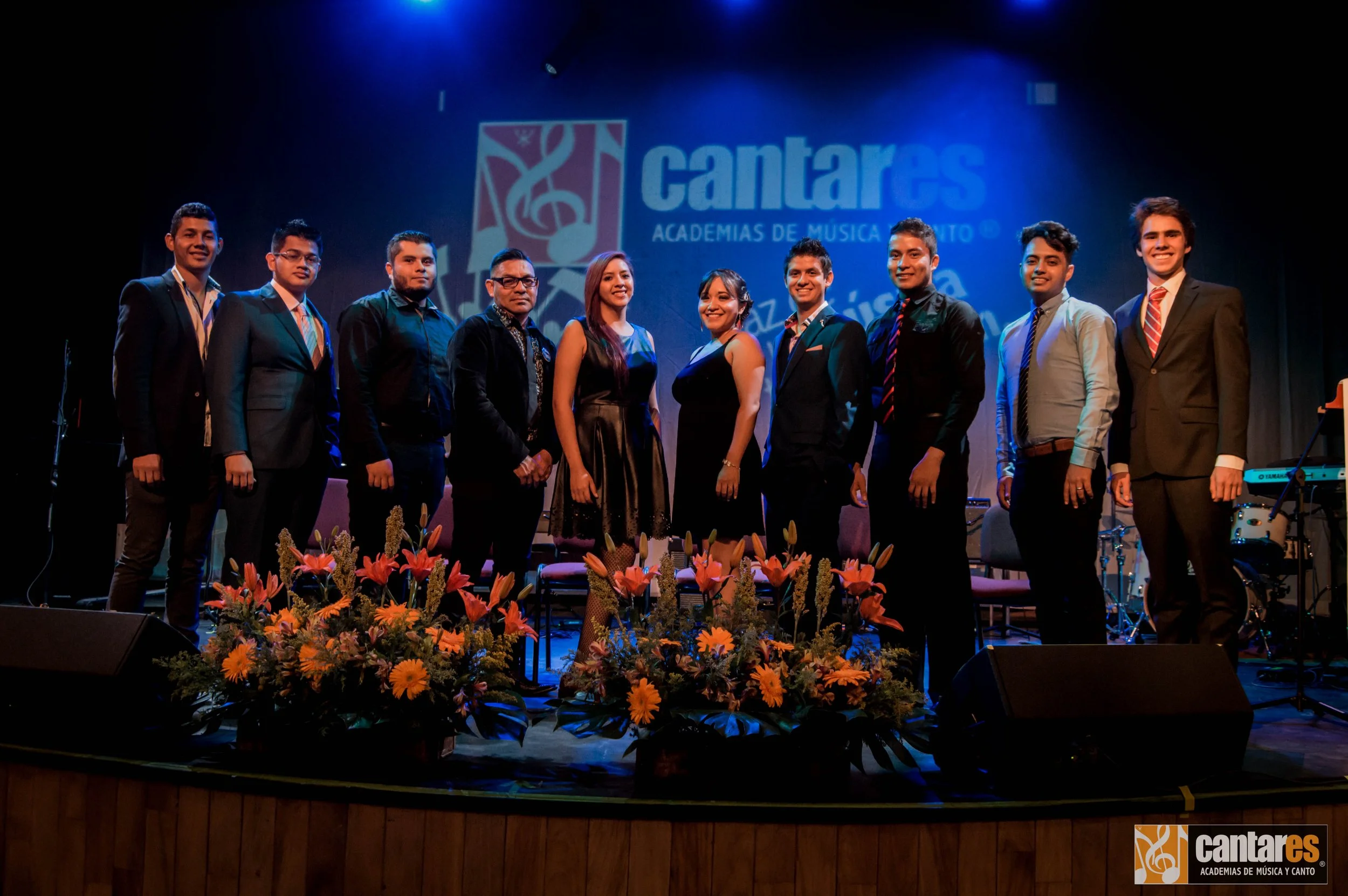 Grupo de varias personas en un escenario con fondo azul que tiene el logo de Cantáres y las palabras "Academias de Música y Canto".
