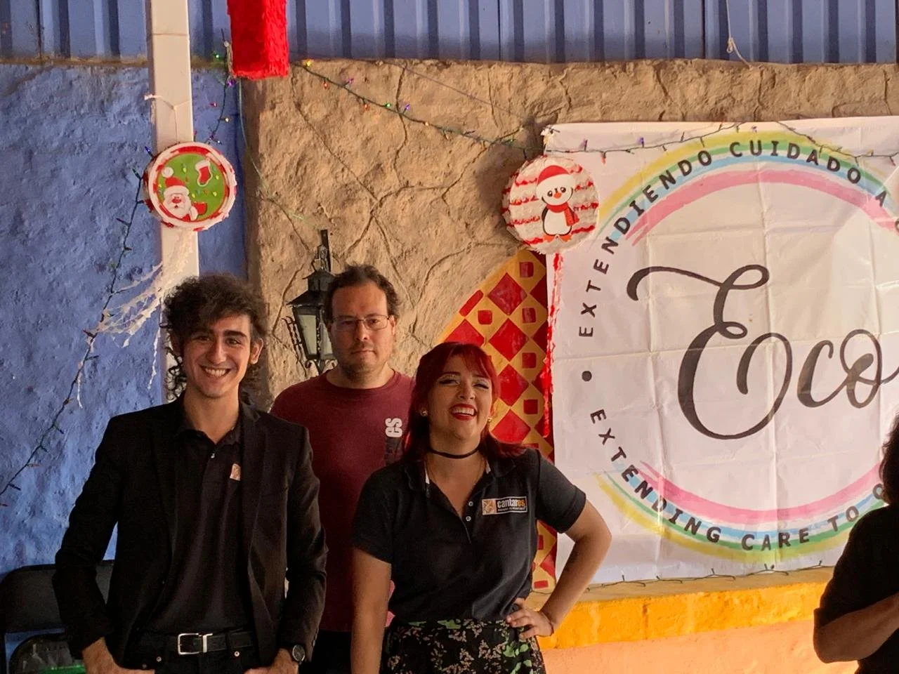 Tres personas posando en una celebración navideña, dos hombres y una mujer, con decoraciones festivas y un cartel con el texto "Extiende tu cuidado".